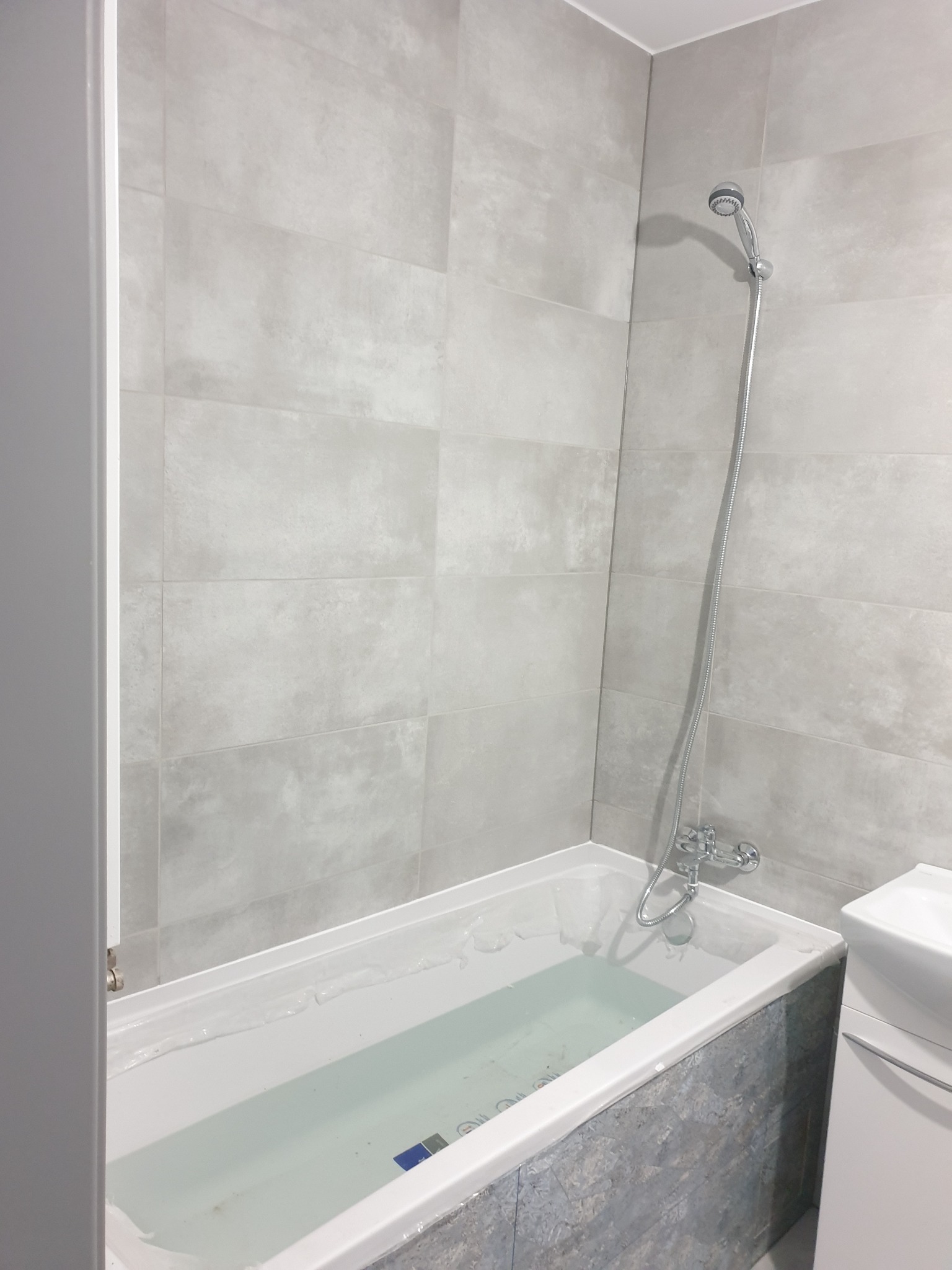 Wypełniona wodą wanna z szarymi, imitującymi kamień panelami dekoracyjnymi, obok umywalka z szafką i ściany wyłożone jasnoszarymi płytkami w dużym formacie.