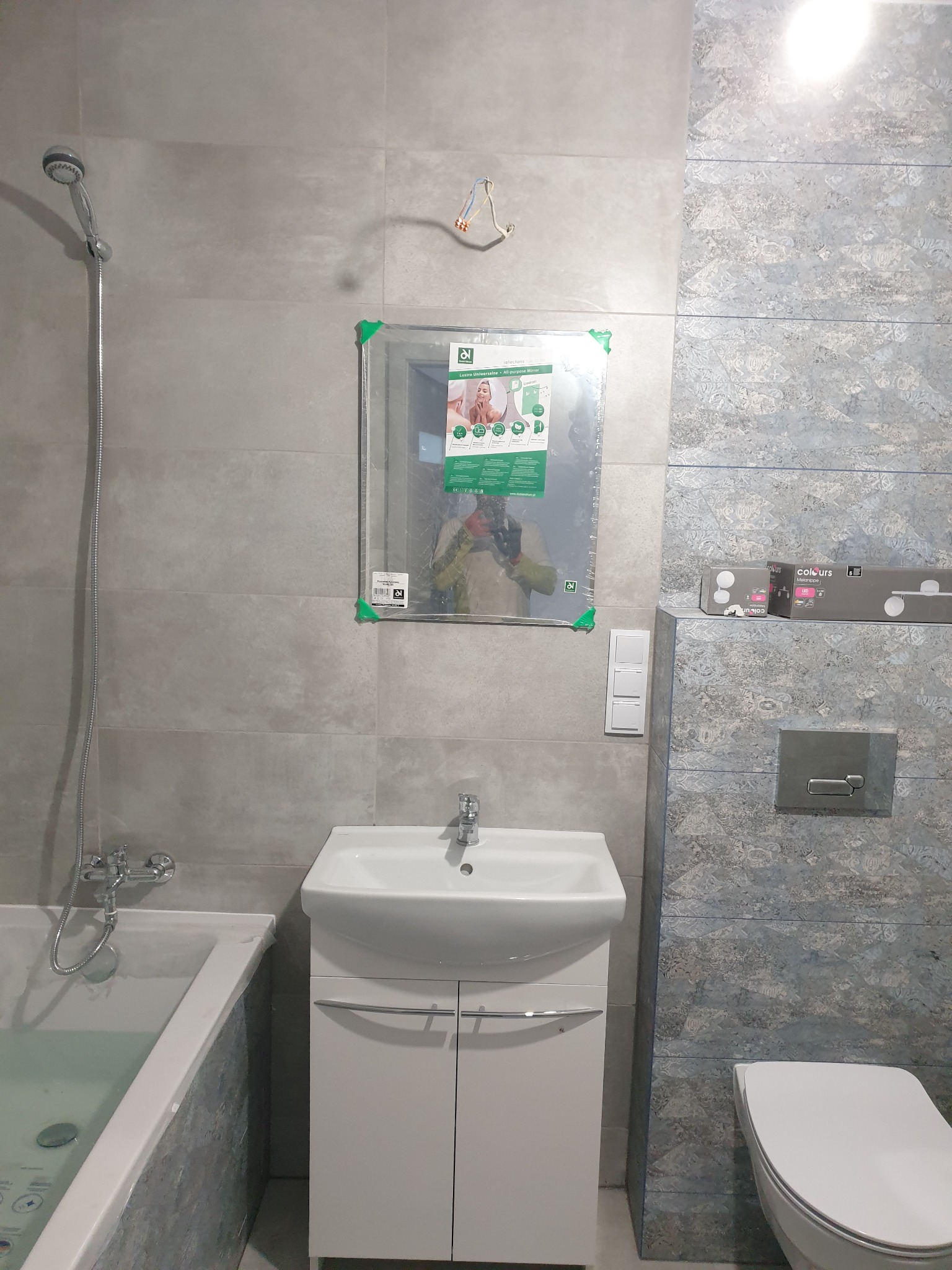 Wykończona łazienka z szarymi płytkami, białą umywalką z szafką, toaletą i wanną. Na ścianie wisi lustro w folii ochronnej i niezidentyfikowane światło w pudełku.