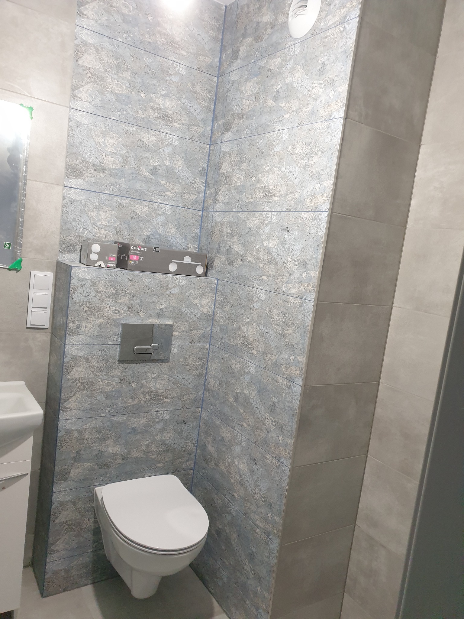 Wykończona łazienka z podwieszaną toaletą, szaro-niebieskie płytki z wzorem, pudełko z oświetleniem LED na obudowie stelaża, widoczny fragment umywalki i panelu sterowania spłuczką.