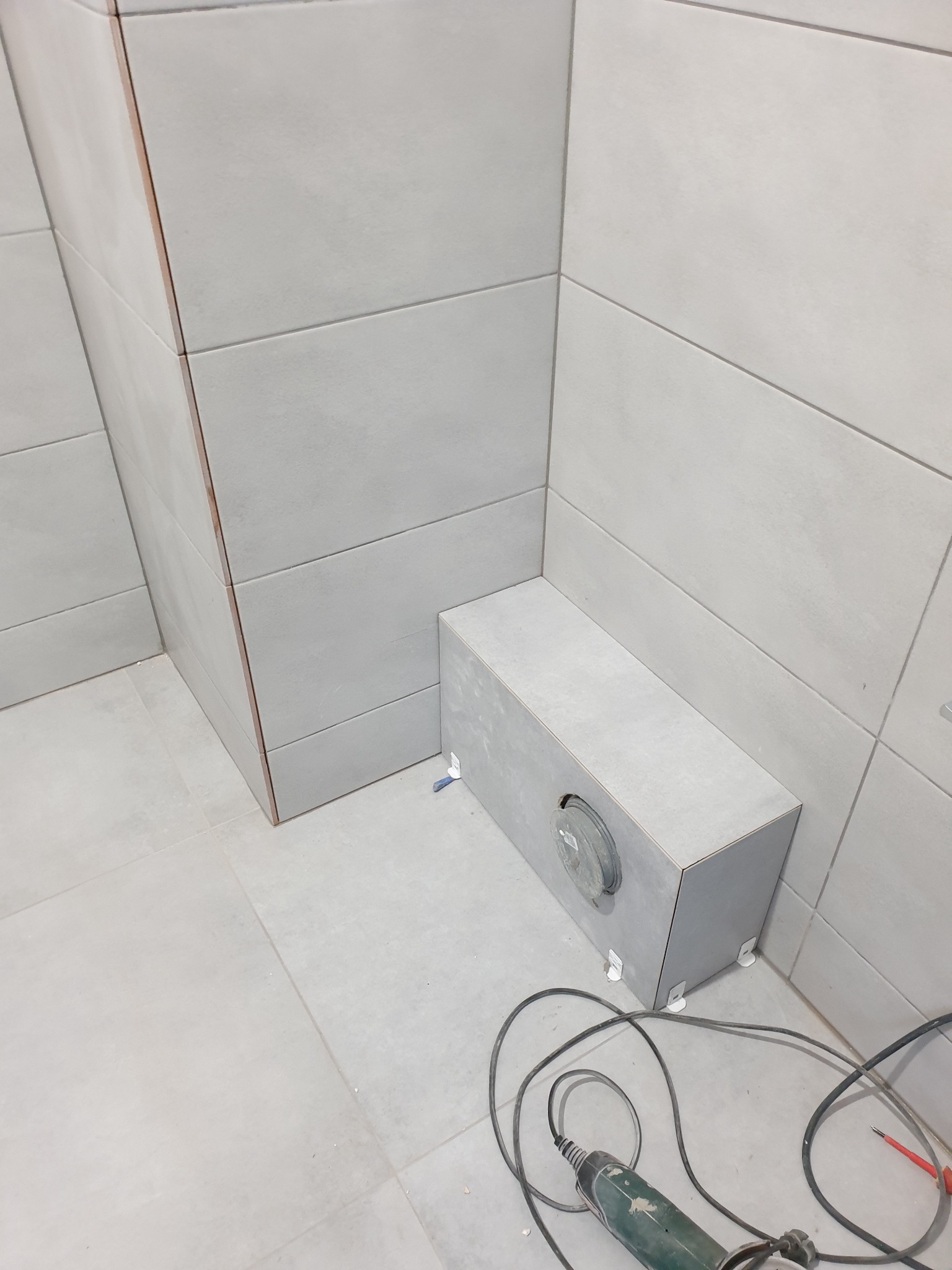 Szara, świeżo wykafelkowana łazienka z widoczną obudową instalacji i narzędziem do cięcia płytek na podłodze.