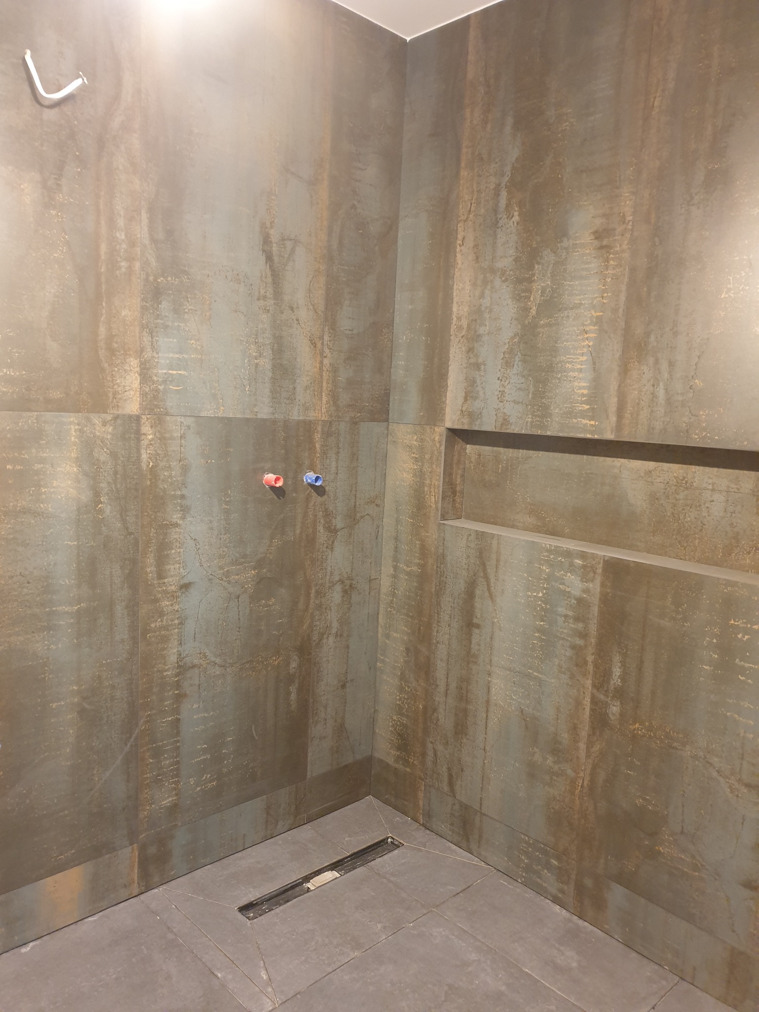 Surowa, industrialna aranżacja kabiny prysznicowej z ciemnymi, metalizowanymi płytkami imitującymi rdzę, widoczne przyłącza wody z zaślepkami i odpływ liniowy w podłodze.