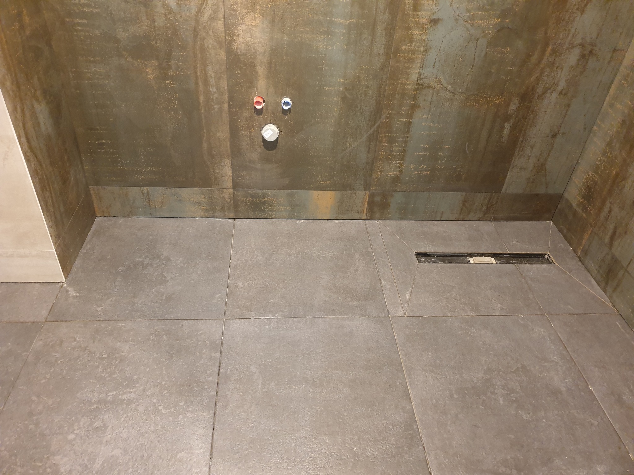 Wykończona łazienka z szarymi płytkami podłogowymi i ścianami imitującymi rdzę, widoczny odpływ liniowy i przyłącza wodne z kolorowymi oznaczeniami.