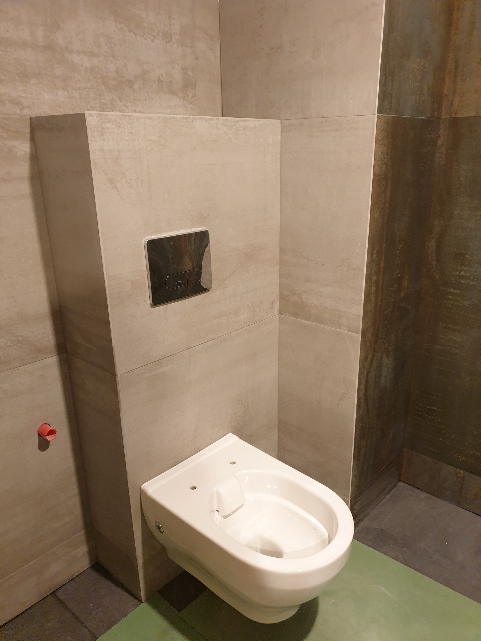 Wisząca miska WC z chromowanym przyciskiem spłukującym, zamontowana na tle płytek imitujących kamień w odcieniach beżu i szarości. Widoczny fragment podłogi z zieloną folią ochronną.