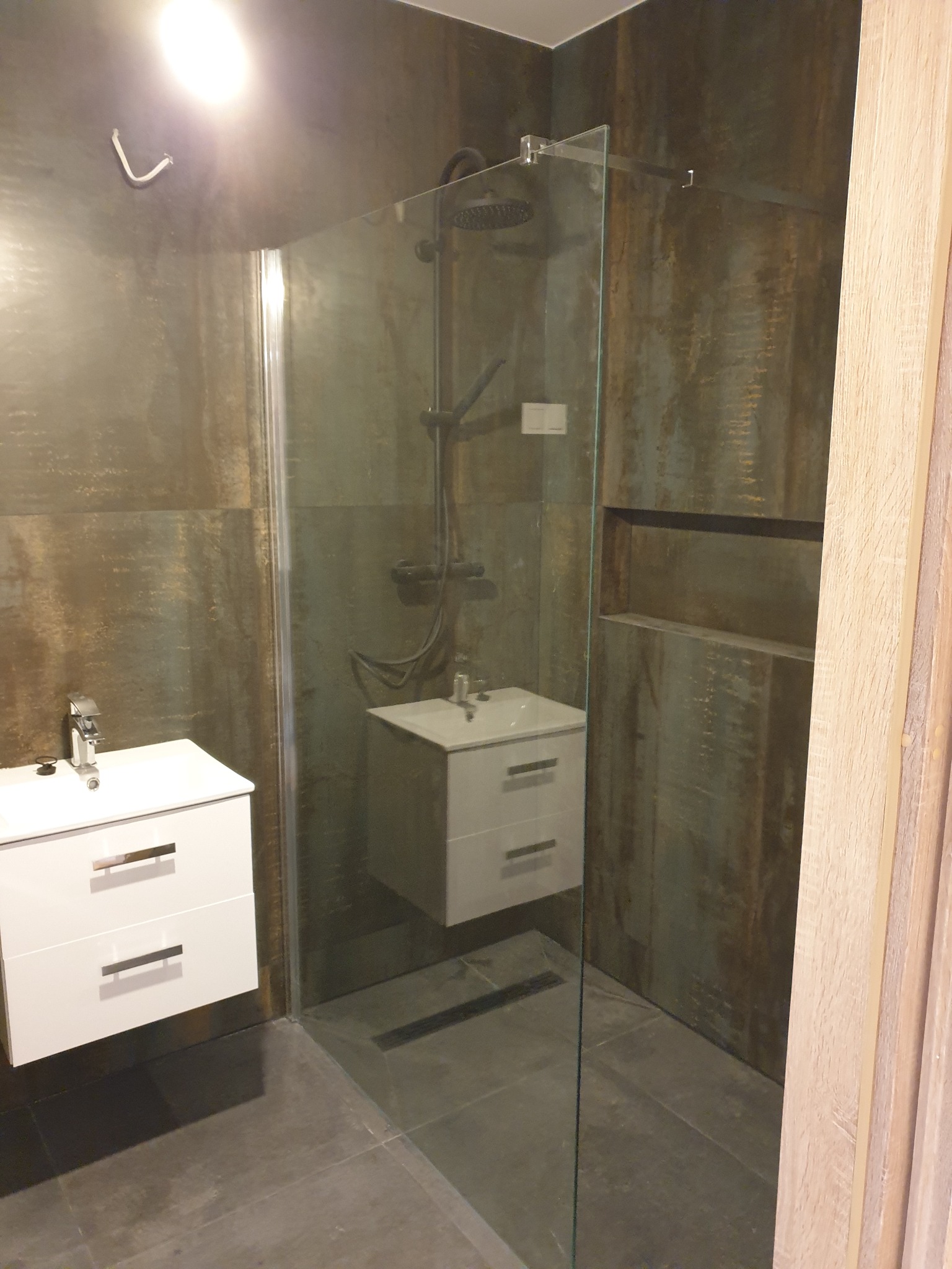 Nowoczesna łazienka z kabiną prysznicową typu walk-in, dwie białe szafki pod umywalki z chromowanymi uchwytami, ściany wykończone płytkami imitującymi rdzę, widoczne mocowania kabiny.