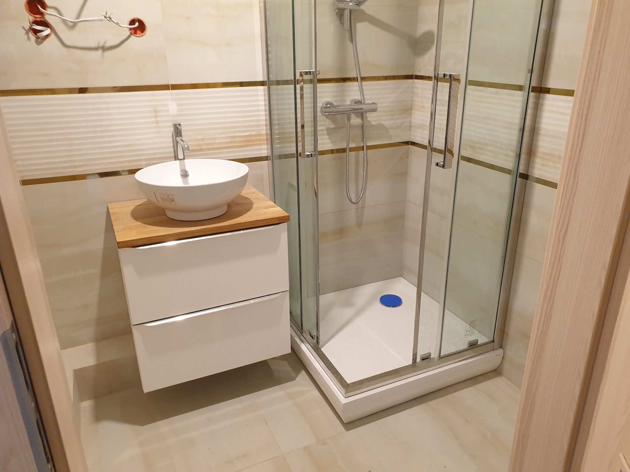 Nowoczesna łazienka po remoncie: biała umywalka nablatowa na drewnianym blacie, białe szafki pod umywalką, kabina prysznicowa z transparentnego szkła, beżowe płytki z poziomymi paskami i złotymi...