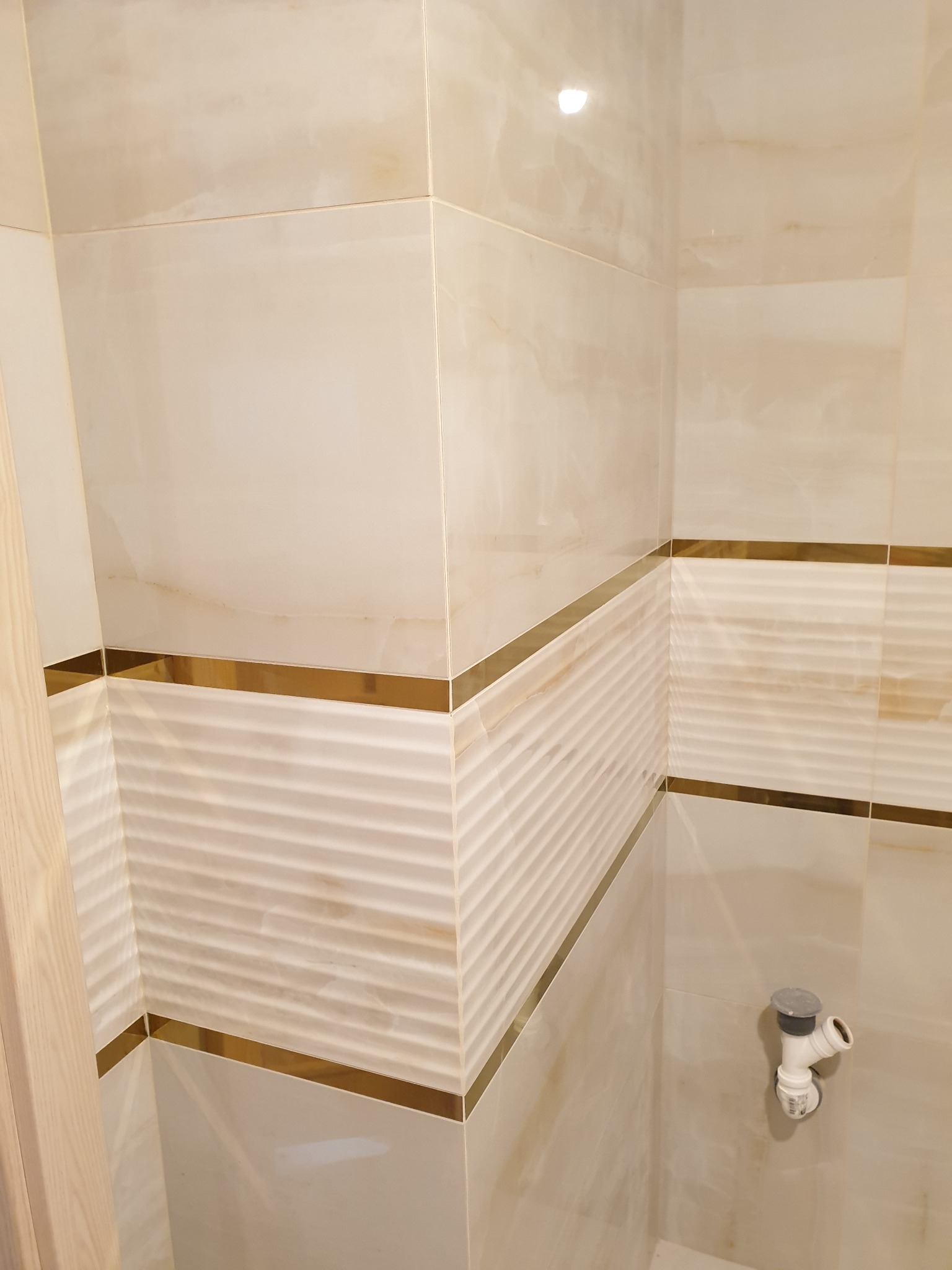 Wykończona łazienka z beżowymi płytkami imitującymi marmur, ozdobiona poziomymi paskami z metalicznym połyskiem i widocznym odpływem pod prysznicem.