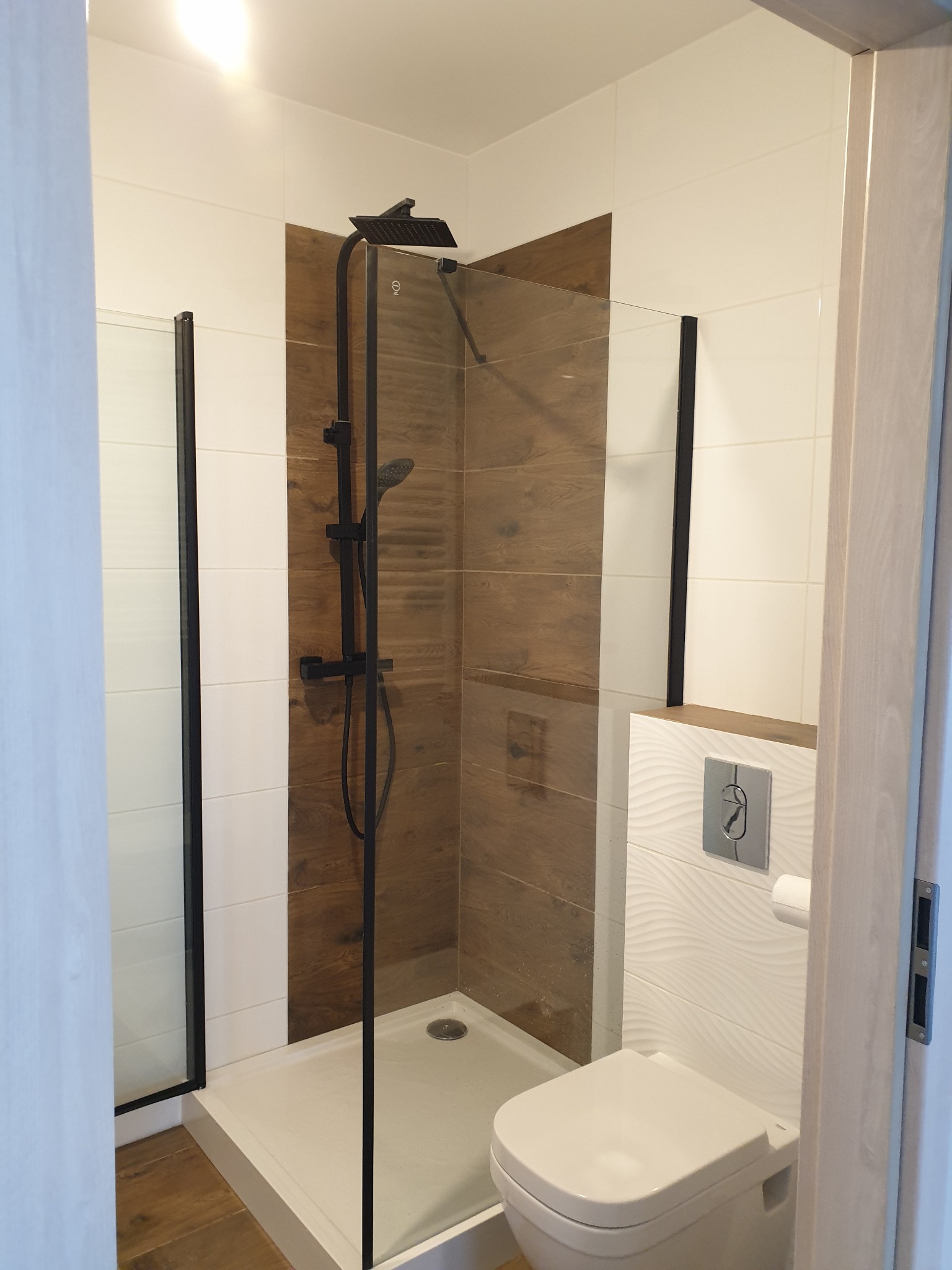 Nowoczesna kabina prysznicowa z czarną armaturą, drewnopodobne płytki na ścianie i podwieszana toaleta z ozdobną płytką falowaną w wykończonej łazience.