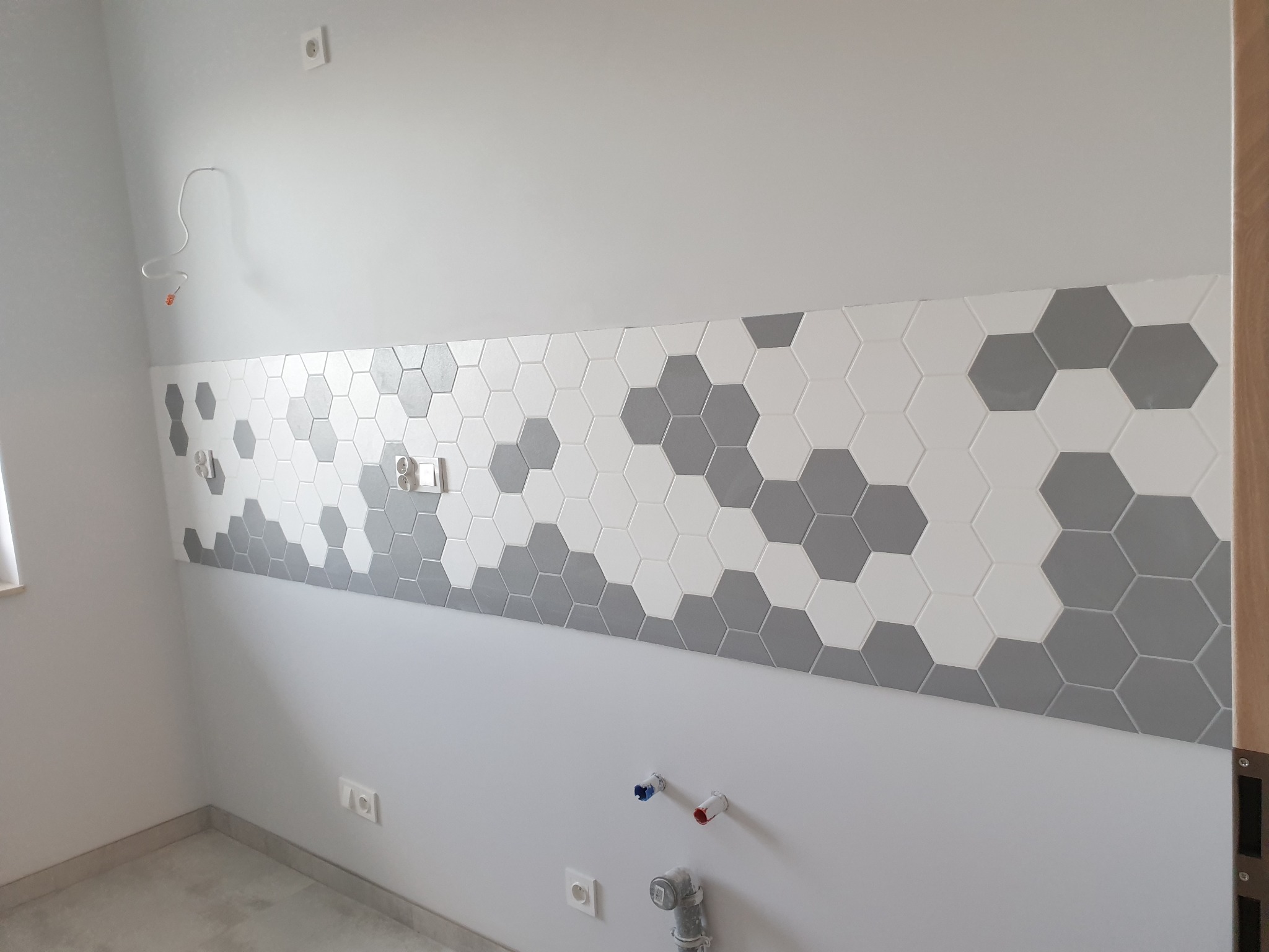Ściana łazienki w trakcie remontu z fragmentem dekoracyjnej mozaiki heksagonalnej w odcieniach szarości i bieli, widoczne wyprowadzenia instalacji wodnej i elektrycznej, surowy stan wykończenia.