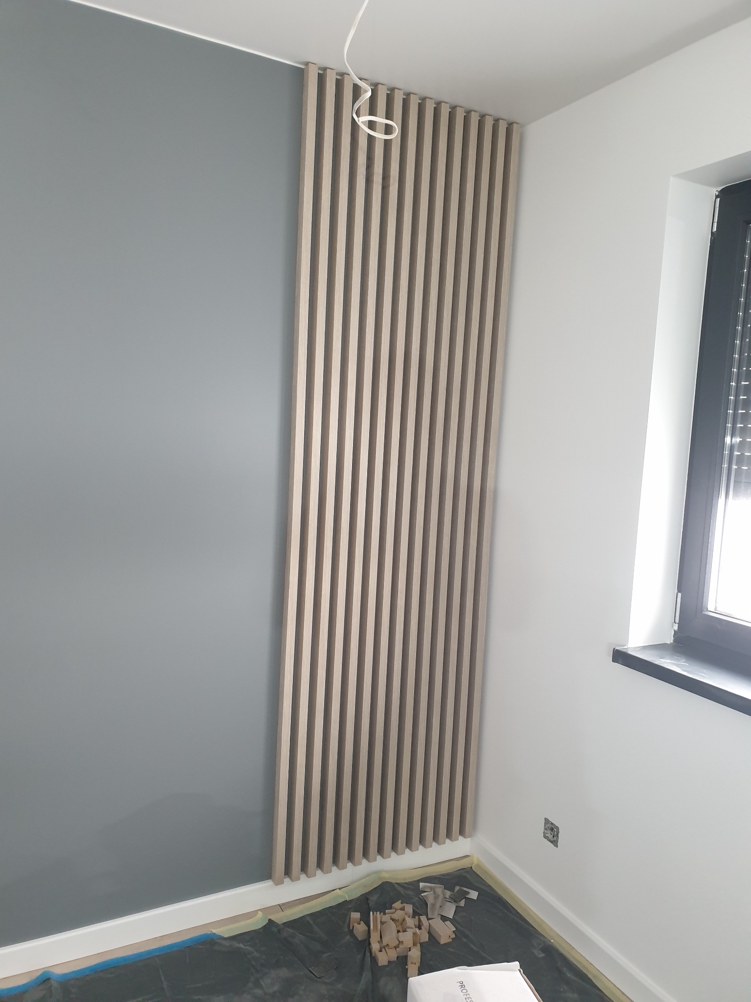 Montaż pionowych, drewnianych lameli dekoracyjnych na szarej ścianie w rogu pokoju, widoczne elementy mocujące i zabezpieczona folią podłoga.