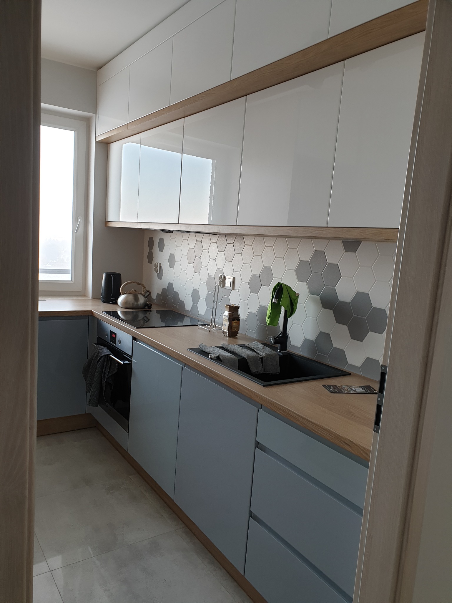 Nowoczesna kuchnia z szarymi szafkami, drewnianym blatem i heksagonalnymi płytkami na ścianie, widok zza framugi drzwi.