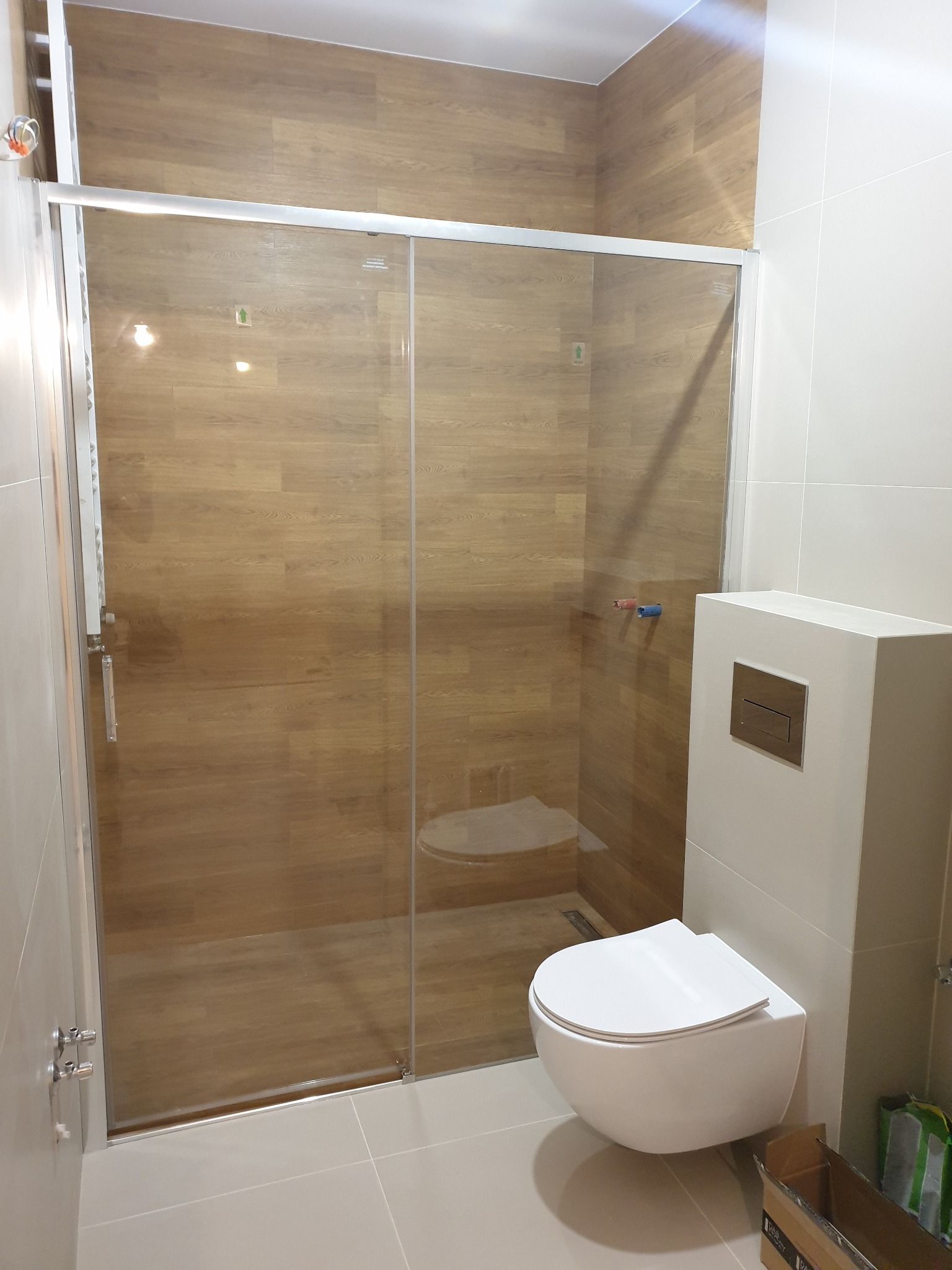 Nowoczesna łazienka z kabiną prysznicową ze szklanymi przesuwnymi drzwiami, podwieszaną toaletą z białym stelażem i płytkami imitującymi drewno na ścianach wewnątrz kabiny.