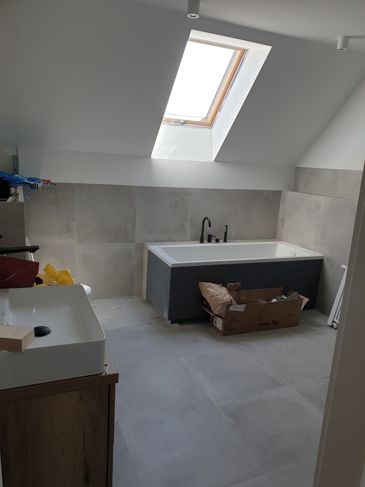 Łazienka w trakcie prac wykończeniowych: zamontowana biała umywalka nablatowa na szafce z drewnopodobnym wykończeniem, obok wanna z czarną armaturą, widoczne szare płytki na ścianach i podłodze...