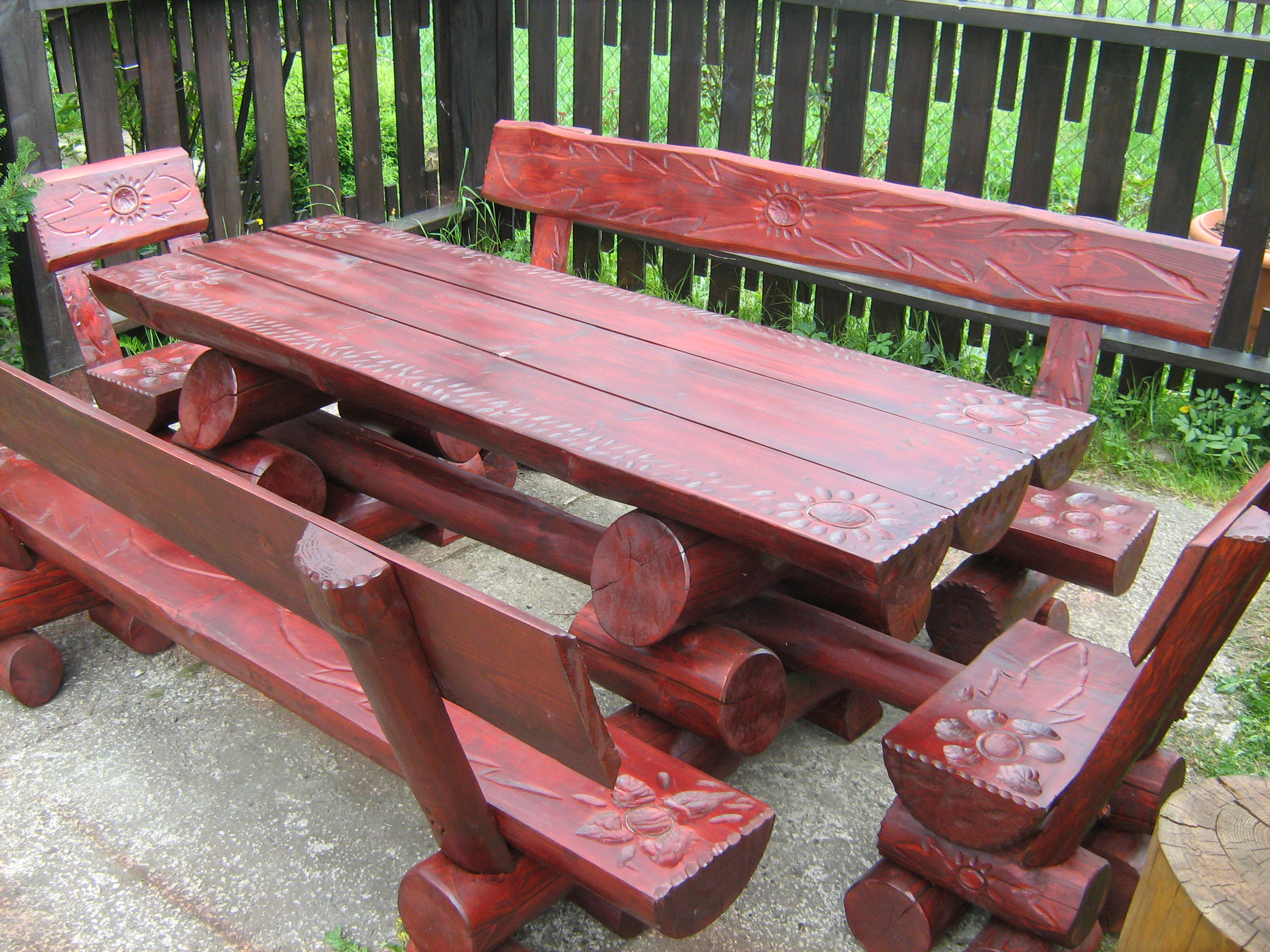 Rustykalny komplet ogrodowy z ciemnego drewna: stół i ławki z oparciami, zdobione motywami kwiatowymi i słonecznymi, ustawiony na betonowym podłożu przed drewnianym płotem.
