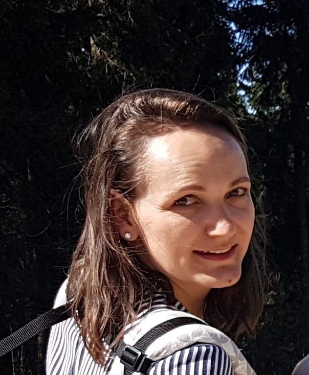 Portret uśmiechniętej kobiety w pasiastym ubraniu, z upiętymi włosami i kolczykiem, na tle zieleni.