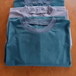 Stos trzech złożonych T-shirtów: dwa turkusowe z szarym ściągaczem i jeden szary z szarym ściągaczem, leżące na drewnianej podłodze.