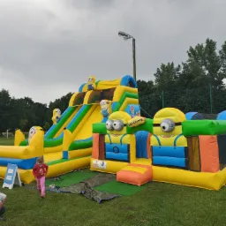 Dmuchany plac zabaw z motywem Minionków na trawie, zjeżdżalnia i tor przeszkód, dziewczynka w różowym ubraniu zbliża się do atrakcji, w tle drzewa i słup oświetleniowy.