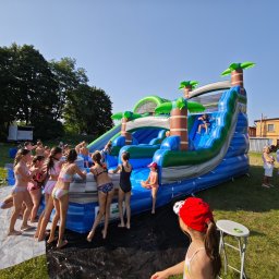 Fun Events Marcin Grysczyk - Dzieci korzystające z dmuchanej zjeżdżalni wodnej z motywem palm na trawiastym terenie podczas słonecznego dnia. W tle budynek i drzewa.