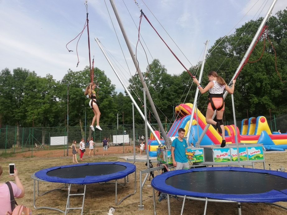 Dwie dziewczynki skaczą na trampolinach bungee, w tle dmuchany park rozrywki i grupa osób obserwujących.