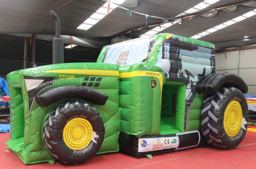 Zielony dmuchany zamek w kształcie traktora John Deere 7310R z czarnymi oponami Michelin i żółtymi felgami, ustawiony w magazynie z widocznymi świetlikami dachowymi.