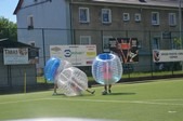 Dwie osoby w przezroczystych kulach do bubble football na zielonym boisku; jedna osoba upada, druga stoi. W tle budynek i reklamy.