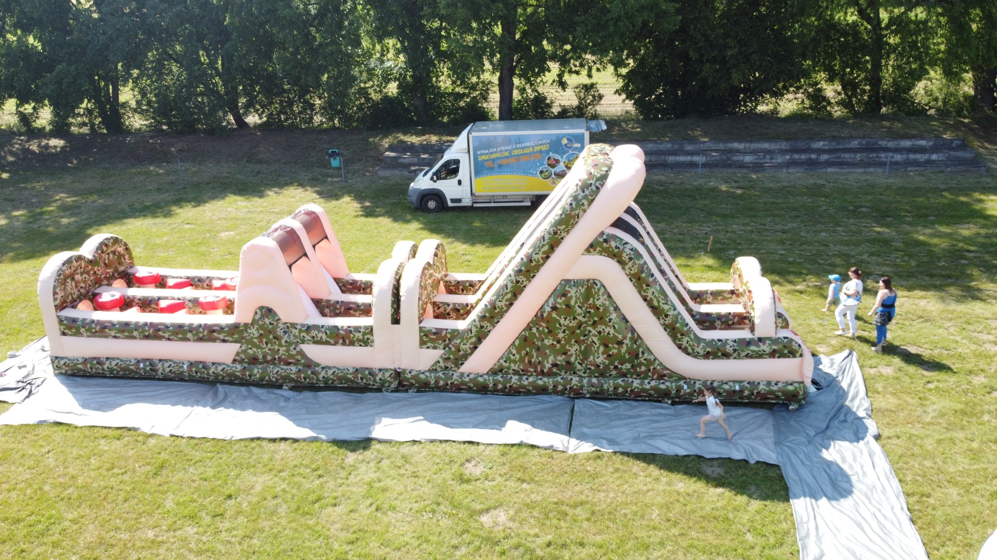 Dmuchany tor przeszkód w kamuflażu na trawie, z widocznym busem z reklamą wynajmu dmuchańców i bawiącymi się dziećmi. Ujęcie z góry, słoneczny dzień.