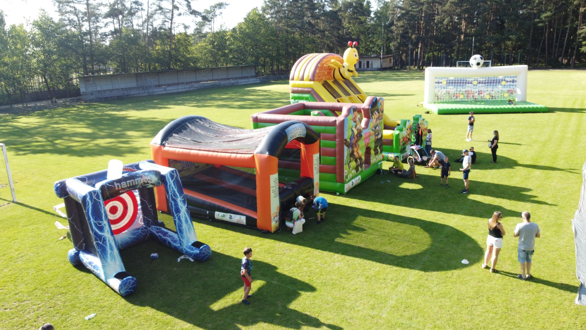 Rozległy teren zielony z rozstawionymi dmuchańcami o różnej tematyce, w tym zjeżdżalnia z pszczołą i bramka do piłki nożnej. Dzieci i dorośli korzystają z atrakcji.