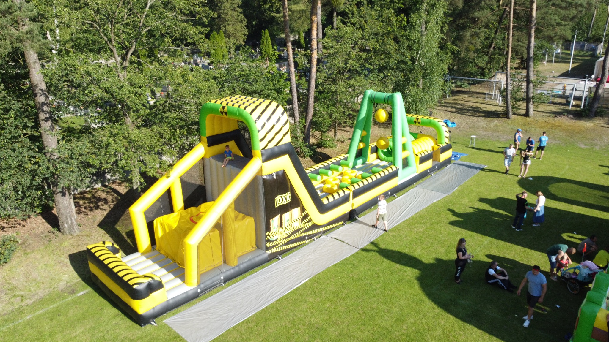 Dmuchany tor przeszkód w czarno-żółtej kolorystyce, z dzieckiem na szczycie, otoczony zielenią i grupą ludzi w tle, uchwycony z lotu ptaka.