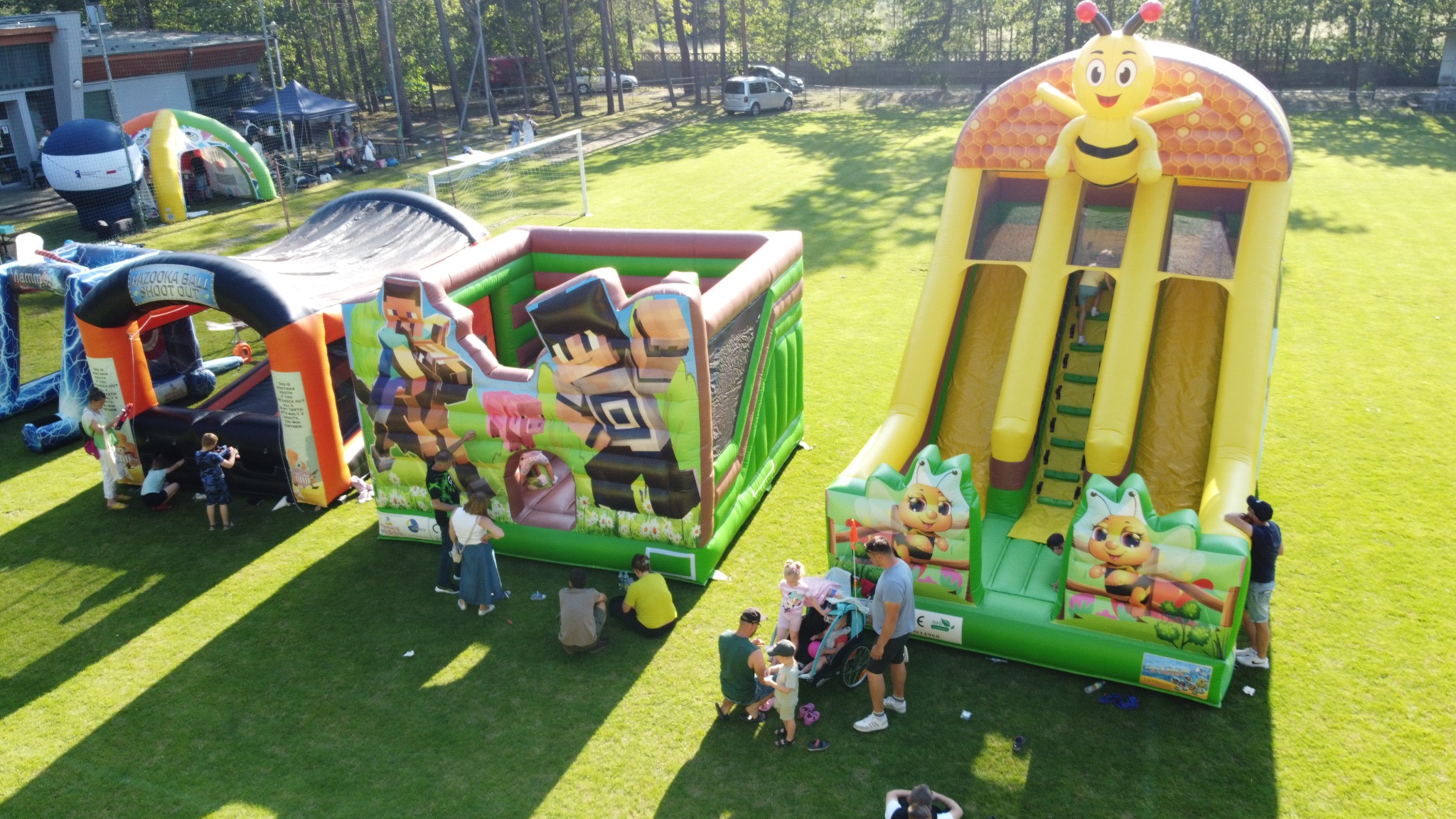 Kolorowe dmuchańce z motywami pszczół i postaci z gier na zielonym trawniku, otoczone bawiącymi się dziećmi i dorosłymi w słoneczny dzień.