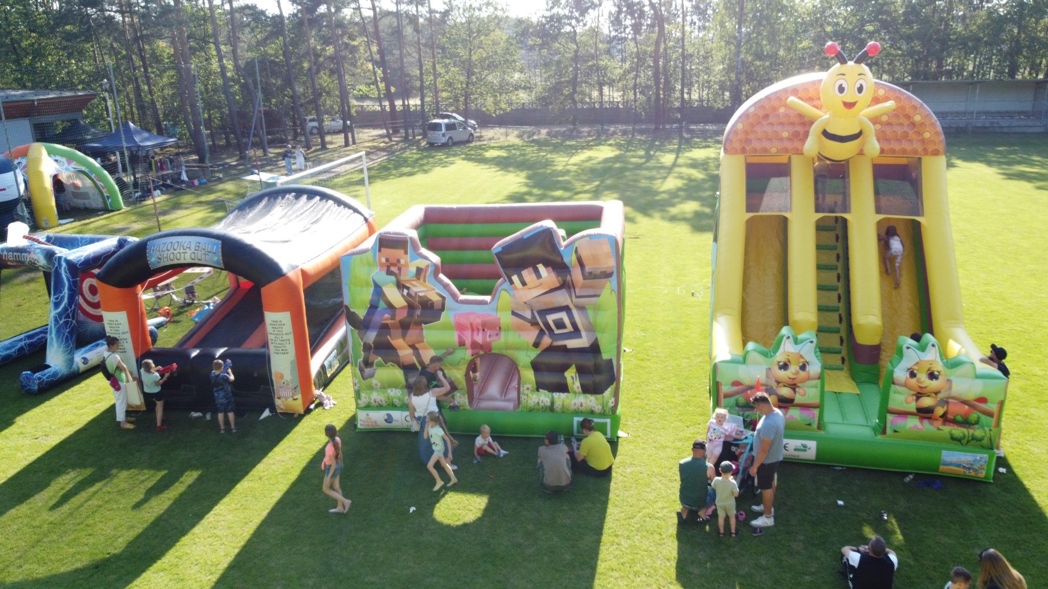 Kolorowe dmuchańce na trawie, w tle las i budynek. Dzieci bawią się na atrakcjach, w tym zjeżdżalnia z motywem pszczoły i dmuchany zamek z postaciami z gier.
