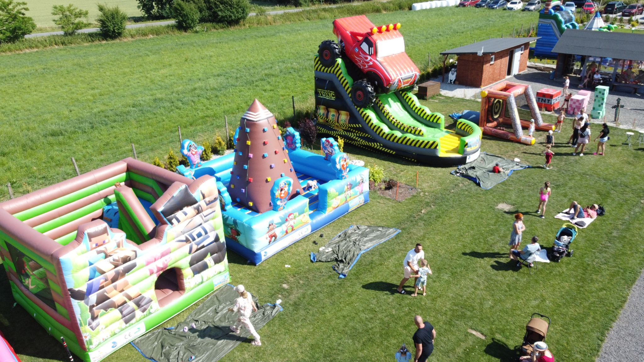 Kolorowe dmuchańce, w tym zjeżdżalnia w kształcie ciężarówki, na zielonym terenie w Opolu. Widoczne osoby korzystające z atrakcji, w tle budynki i zaparkowane samochody.