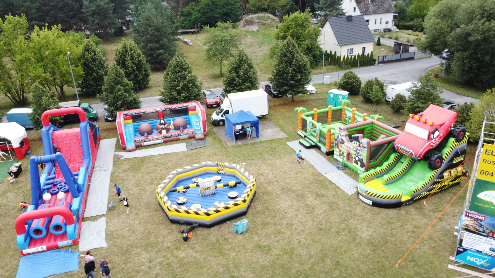 Widok z góry na rozstawione dmuchańce: zjeżdżalnia w kształcie ciężarówki, tor przeszkód, arena z przeszkodami, zamek. W tle drzewa i zabudowania.
