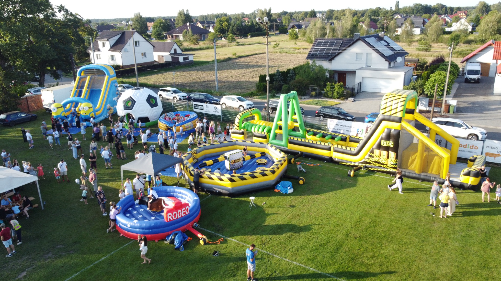 Z lotu ptaka: rozstawione dmuchańce (zjeżdżalnia Minionki, rodeo, tor przeszkód) na trawniku podczas imprezy plenerowej z udziałem dużej grupy osób.