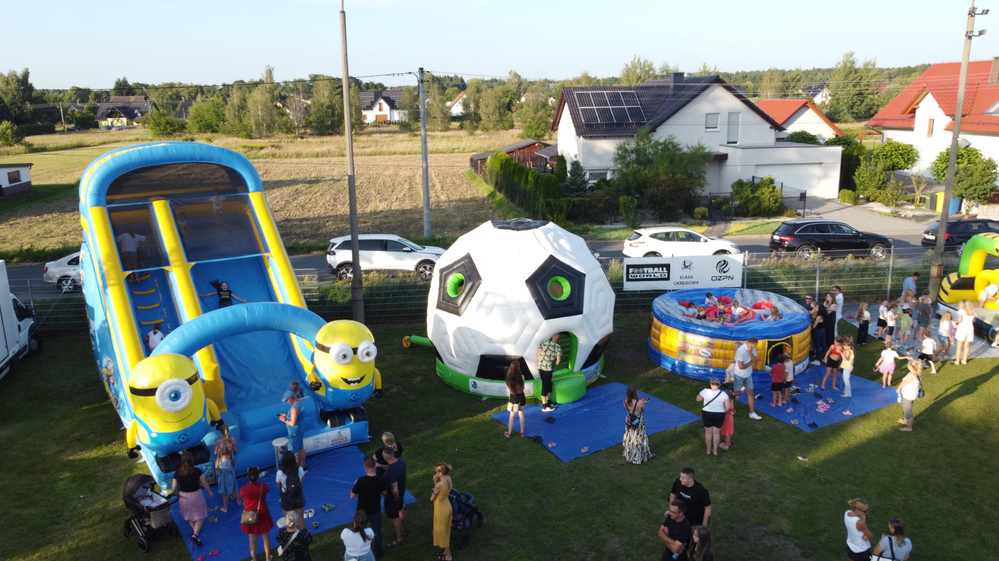 Kompleks dmuchańców: zjeżdżalnia z Minionkami, piłka nożna i basen z piłeczkami. Dzieci i dorośli bawią się na trawie w słoneczny dzień. Widok z góry.