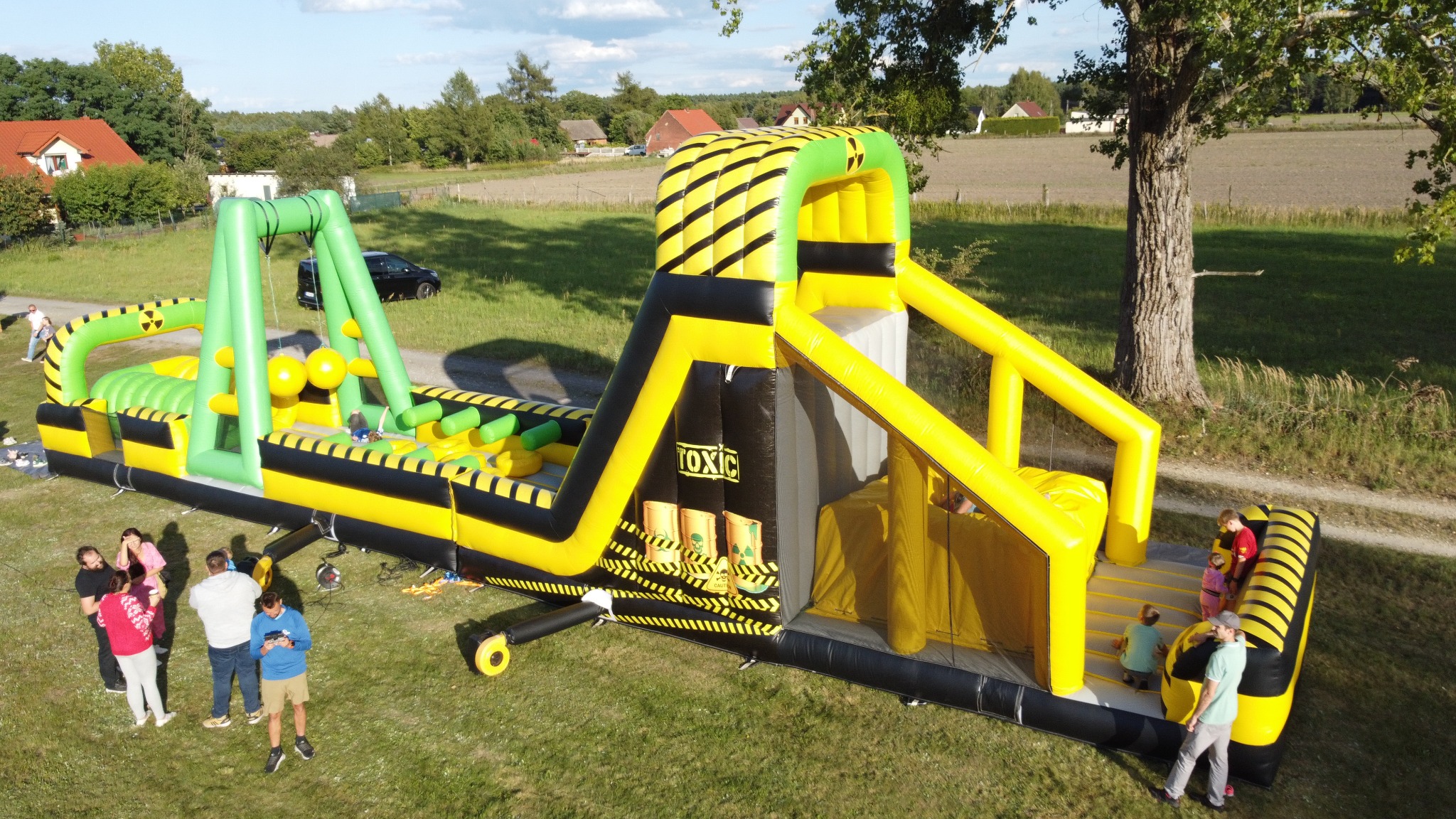 Żółto-czarny dmuchany tor przeszkód z motywem ostrzegawczym 'Toxic' na trawie, z bawiącymi się dziećmi i grupą dorosłych w tle, w słoneczny dzień.