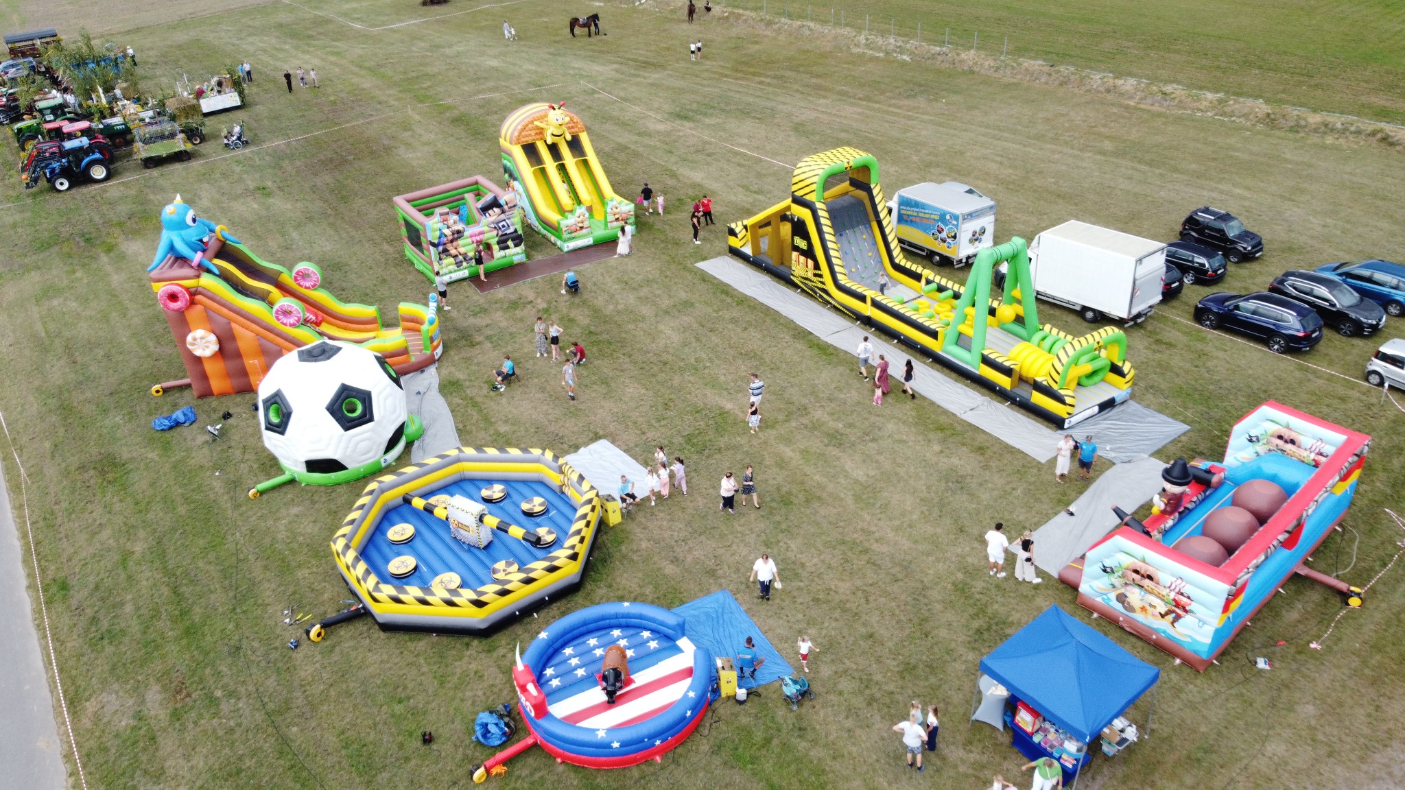 Widok z góry na rozstawione dmuchańce na trawiastym terenie, z bawiącymi się dziećmi i dorosłymi. W tle widoczne pojazdy i drzewa.