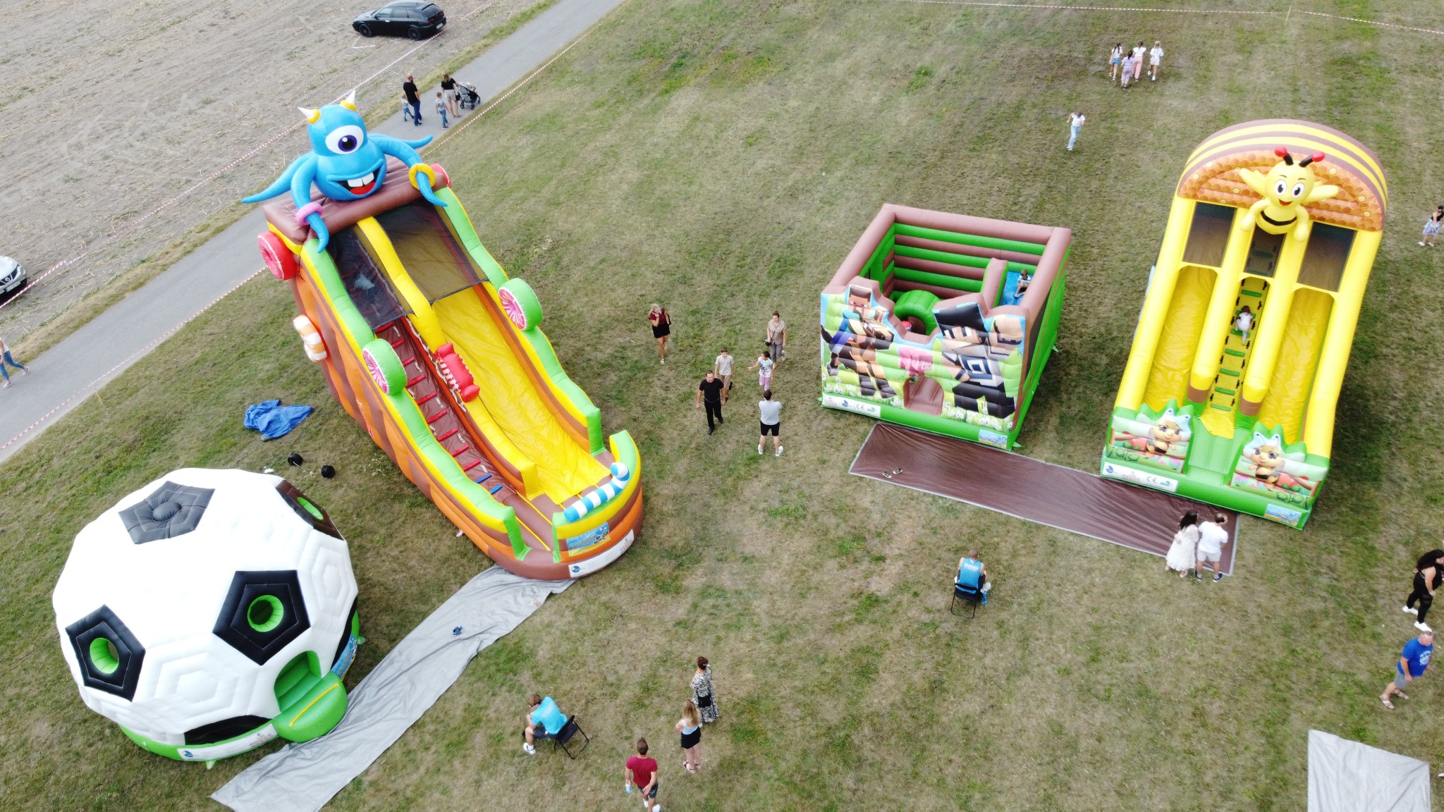 Widok z góry na rozstawione dmuchańce: zjeżdżalnia z potworem, kwadratowy zamek i zjeżdżalnia z pszczołą. Obok dmuchana piłka. Ludzie w tle. Letni dzień.