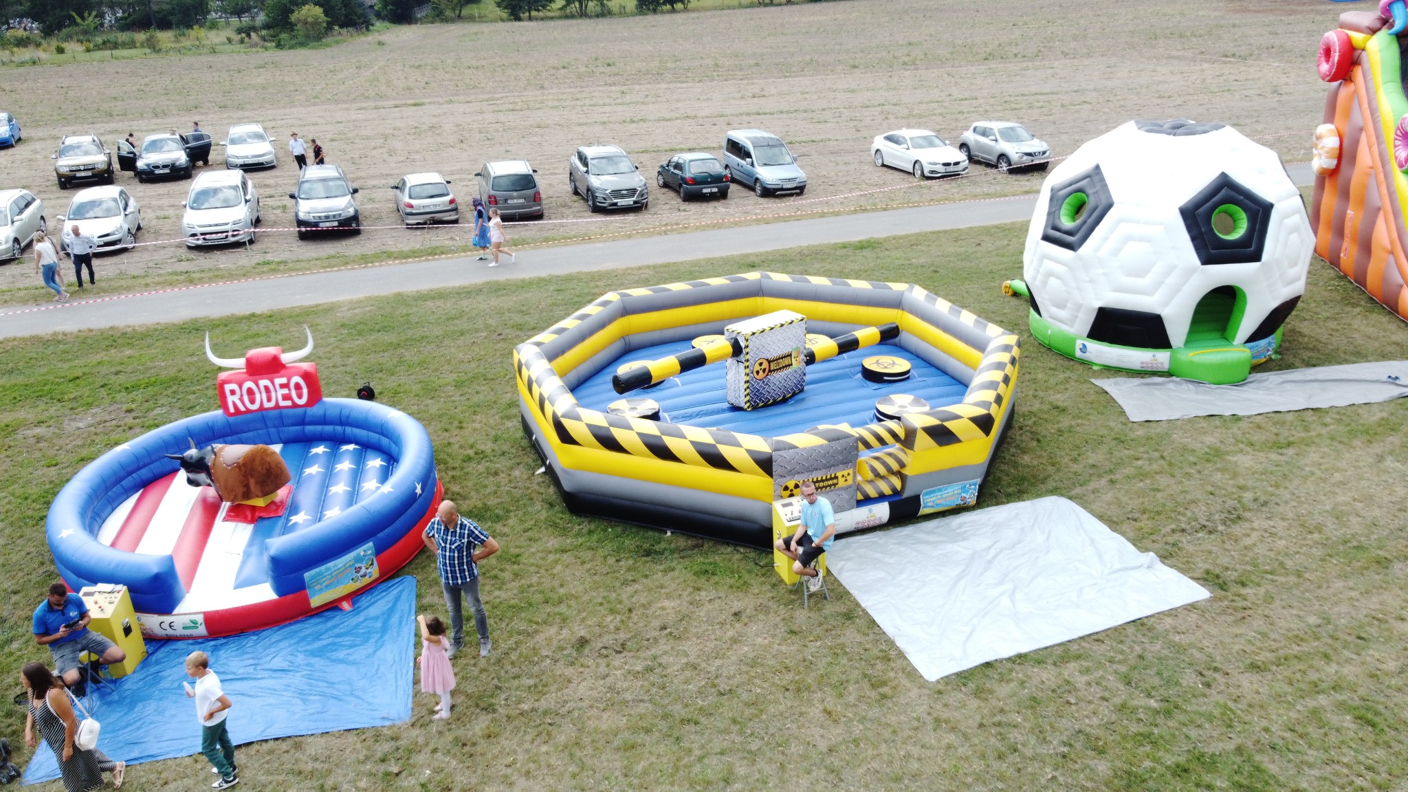 Z lotu ptaka: rozstawione dmuchańce (byk rodeo, wipeout, piłka nożna) na trawie, w tle zaparkowane samochody i spacerujący ludzie. Impreza plenerowa.