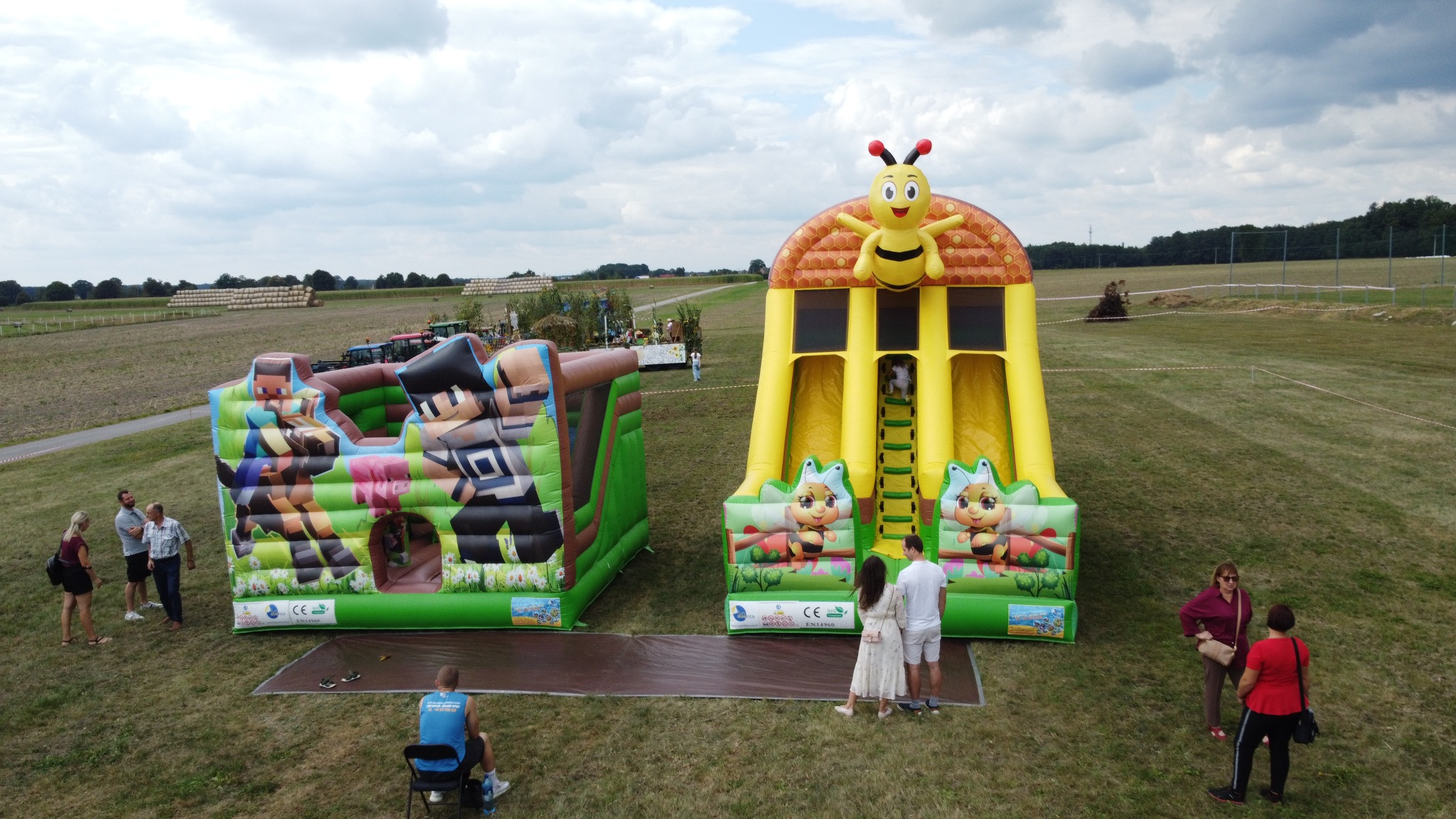 Dwa dmuchańce: Minecraft i pszczoła, na trawiastym terenie z ludźmi. Widok z góry, w tle pole i las. Dmuchańce na wynajem.