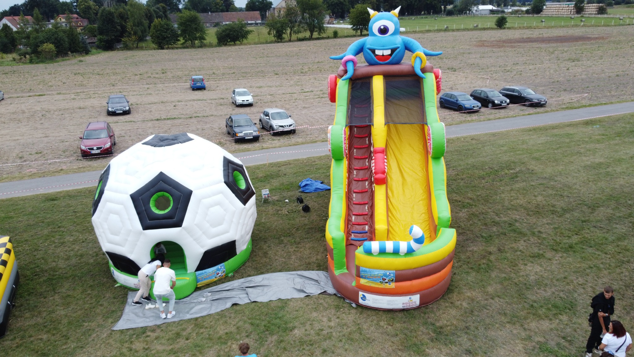 Dwa dmuchańce: zjeżdżalnia z potworem na szczycie i piłka nożna z wejściem, na trawiastym terenie z zaparkowanymi samochodami w tle.
