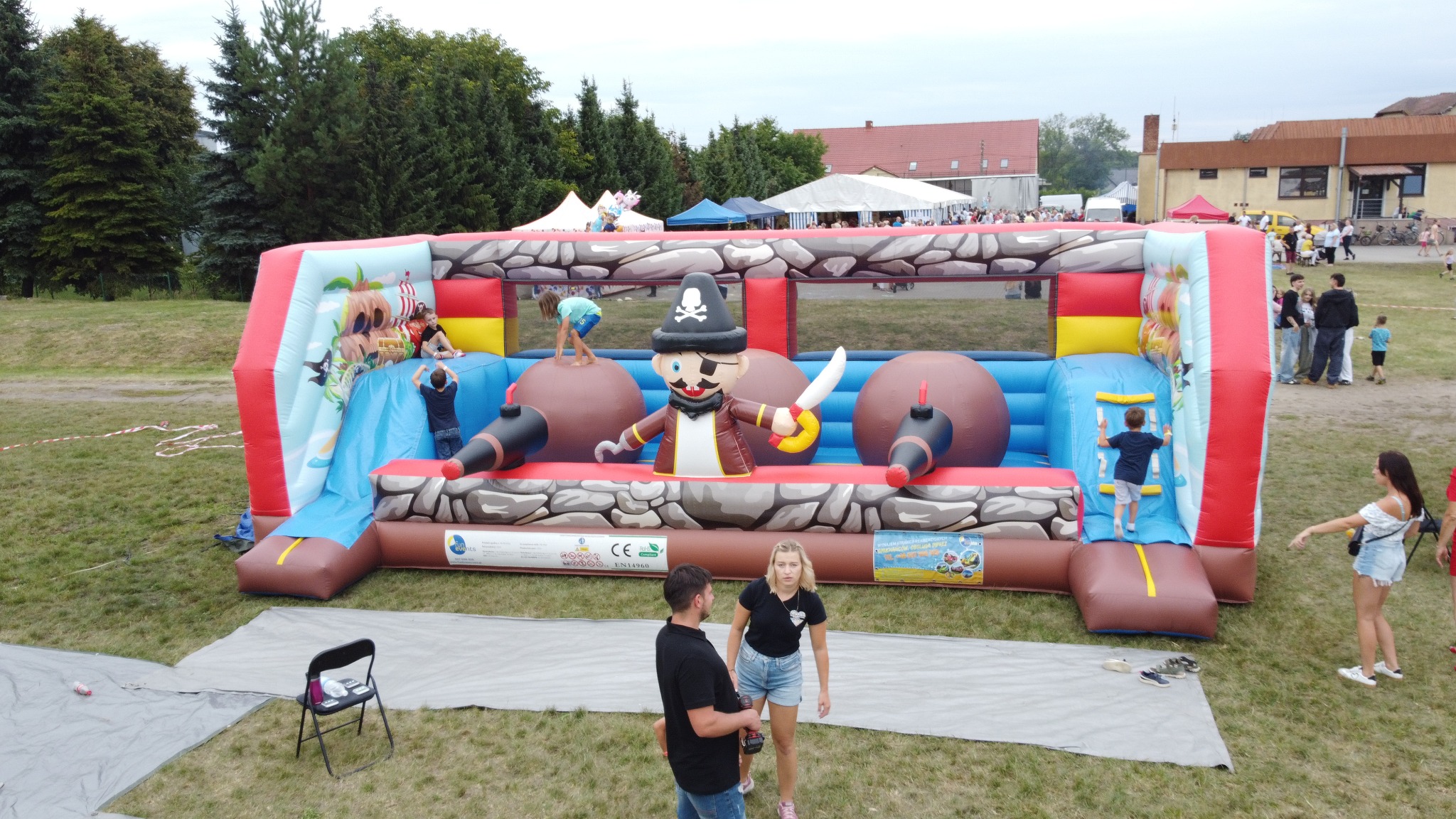 Duży, kolorowy dmuchany zamek w stylu pirackim z figurą pirata. Dzieci bawią się na zjeżdżalni i wewnątrz konstrukcji. W tle widoczni ludzie i budynki.