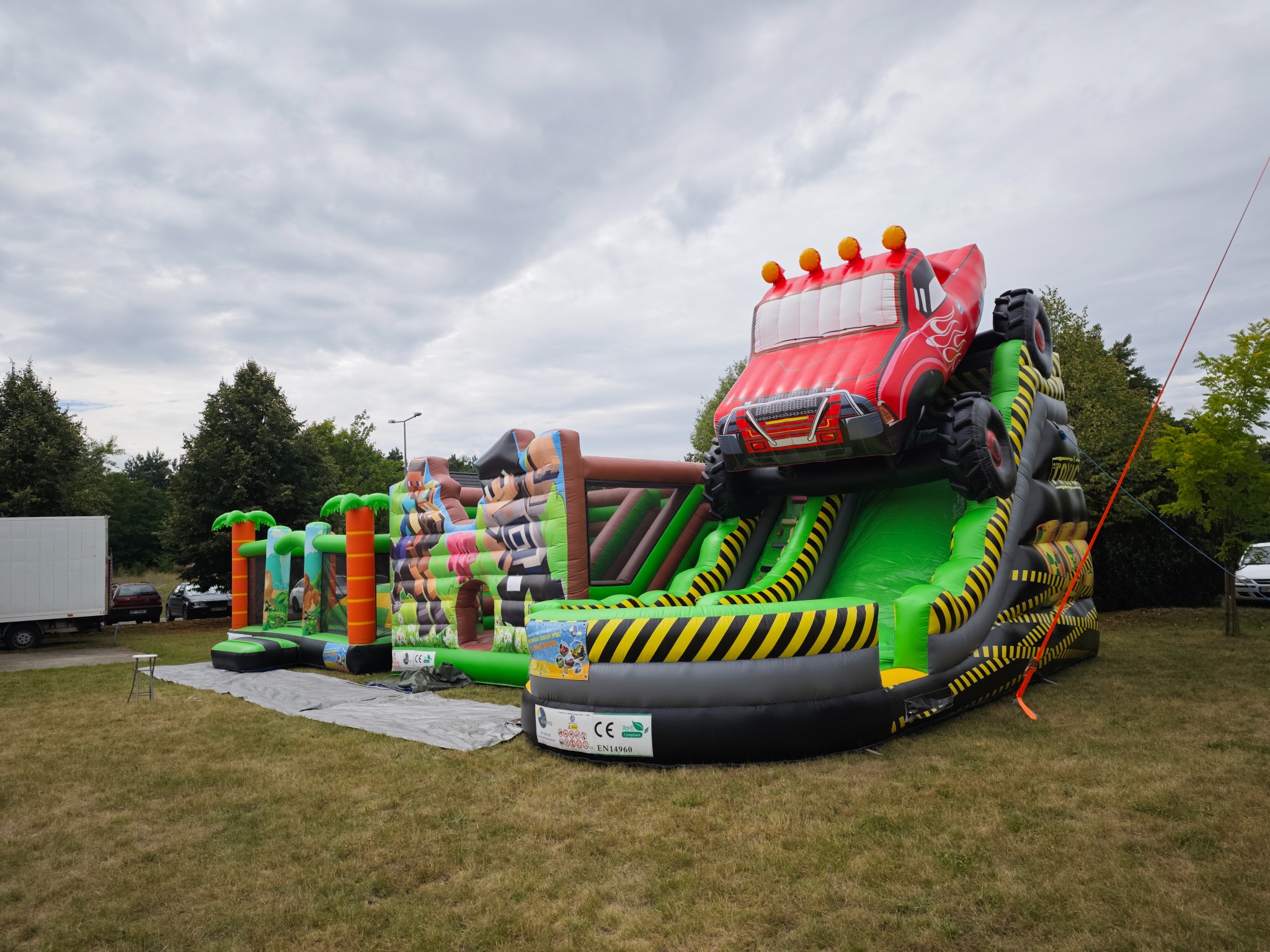 Rozłożony, kolorowy dmuchany zamek z motywem monster trucka na trawie, gotowy do zabawy. Widoczne drzewa i pochmurne niebo w tle.