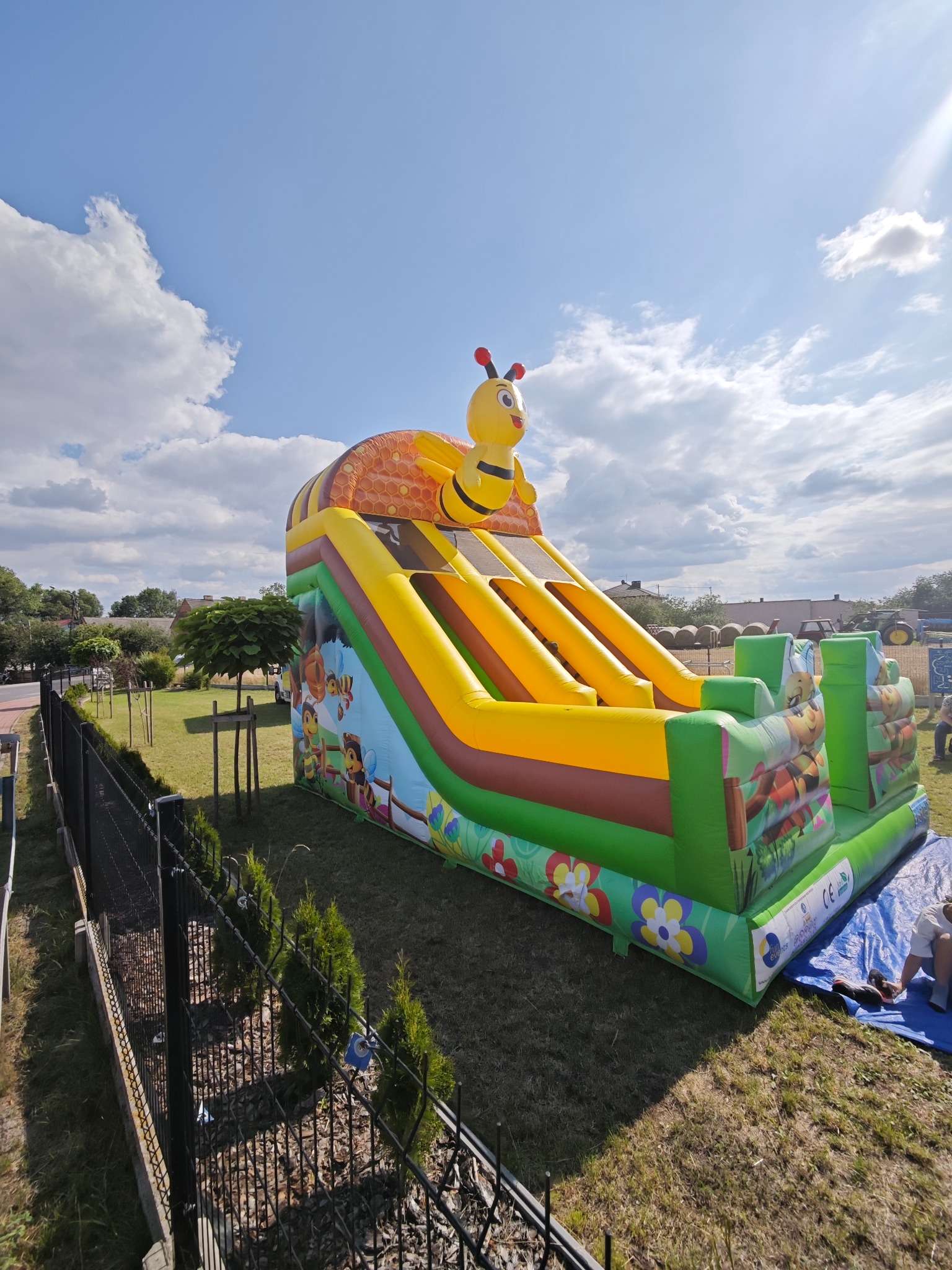 Kolorowa, dmuchana zjeżdżalnia z motywem pszczoły na trawie, widok z dołu, błękitne niebo z chmurami w tle. Zabezpieczona siatką.