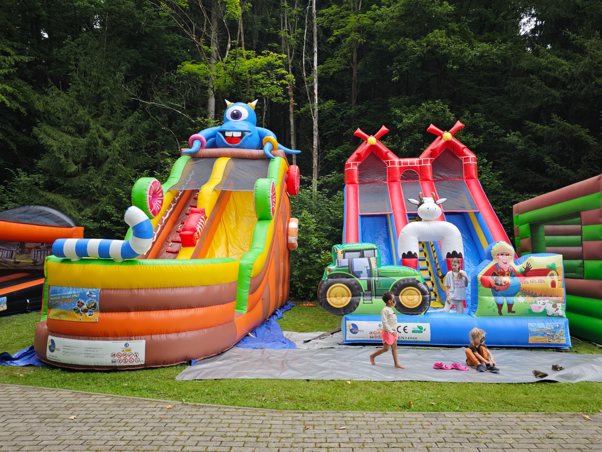 Dwa kolorowe dmuchańce na trawie: jeden z potworem, drugi z traktorem i wiejską scenerią. Dzieci bawią się w pobliżu, a w tle widać drzewa.