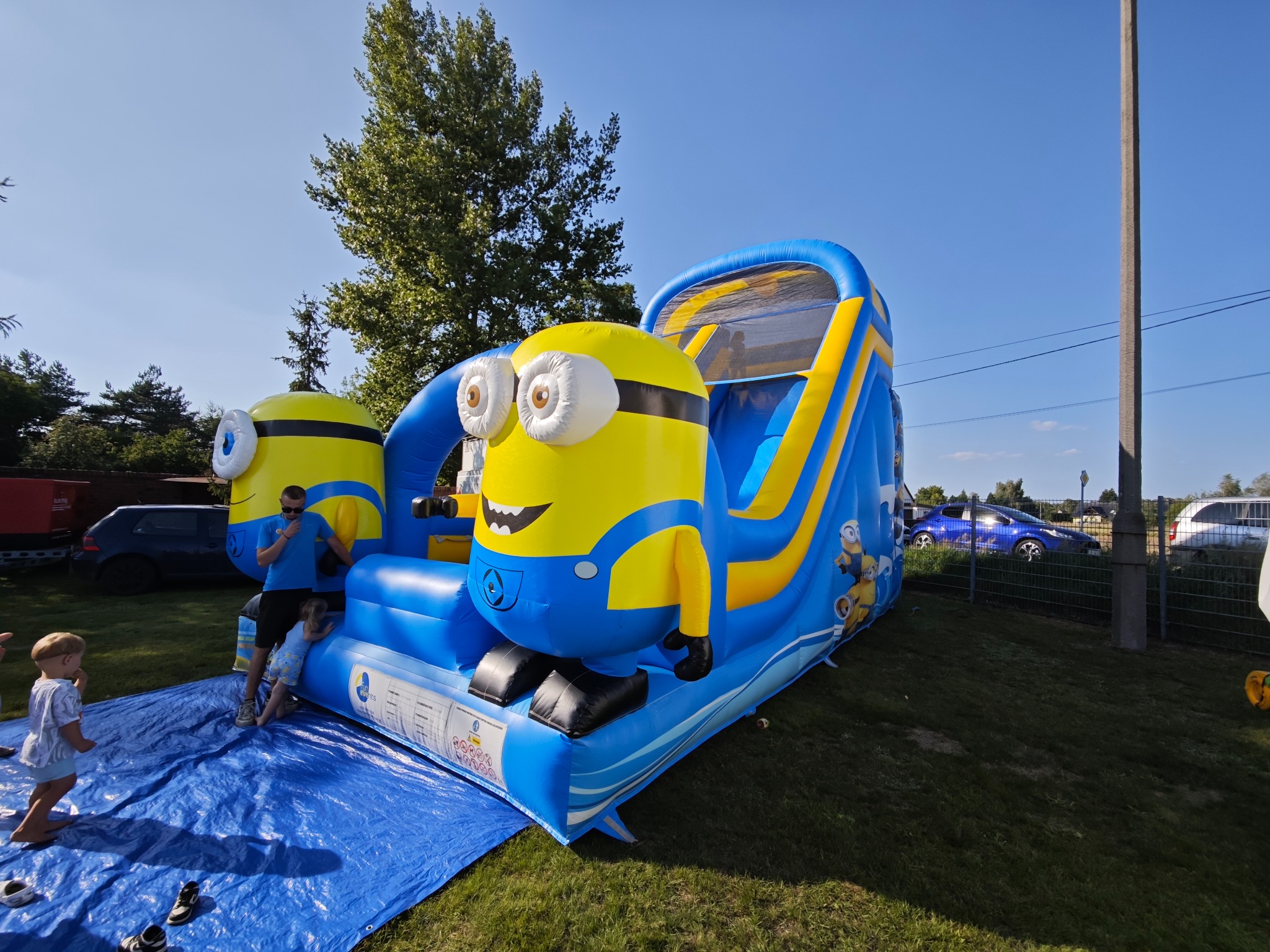 Niebiesko-żółty dmuchany zamek ze zjeżdżalnią w kształcie Minionka, dzieci i dorosły w pobliżu, rozłożona plandeka, trawa, drzewa i samochody w tle.