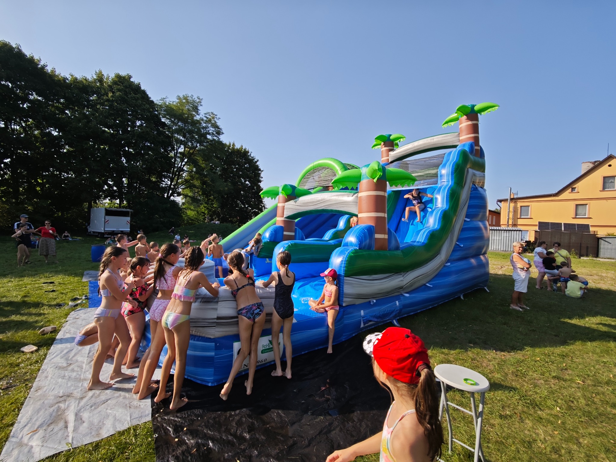 Dzieci korzystające z dmuchanej zjeżdżalni wodnej z motywem palm na trawiastym terenie podczas słonecznego dnia. W tle budynek i drzewa.
