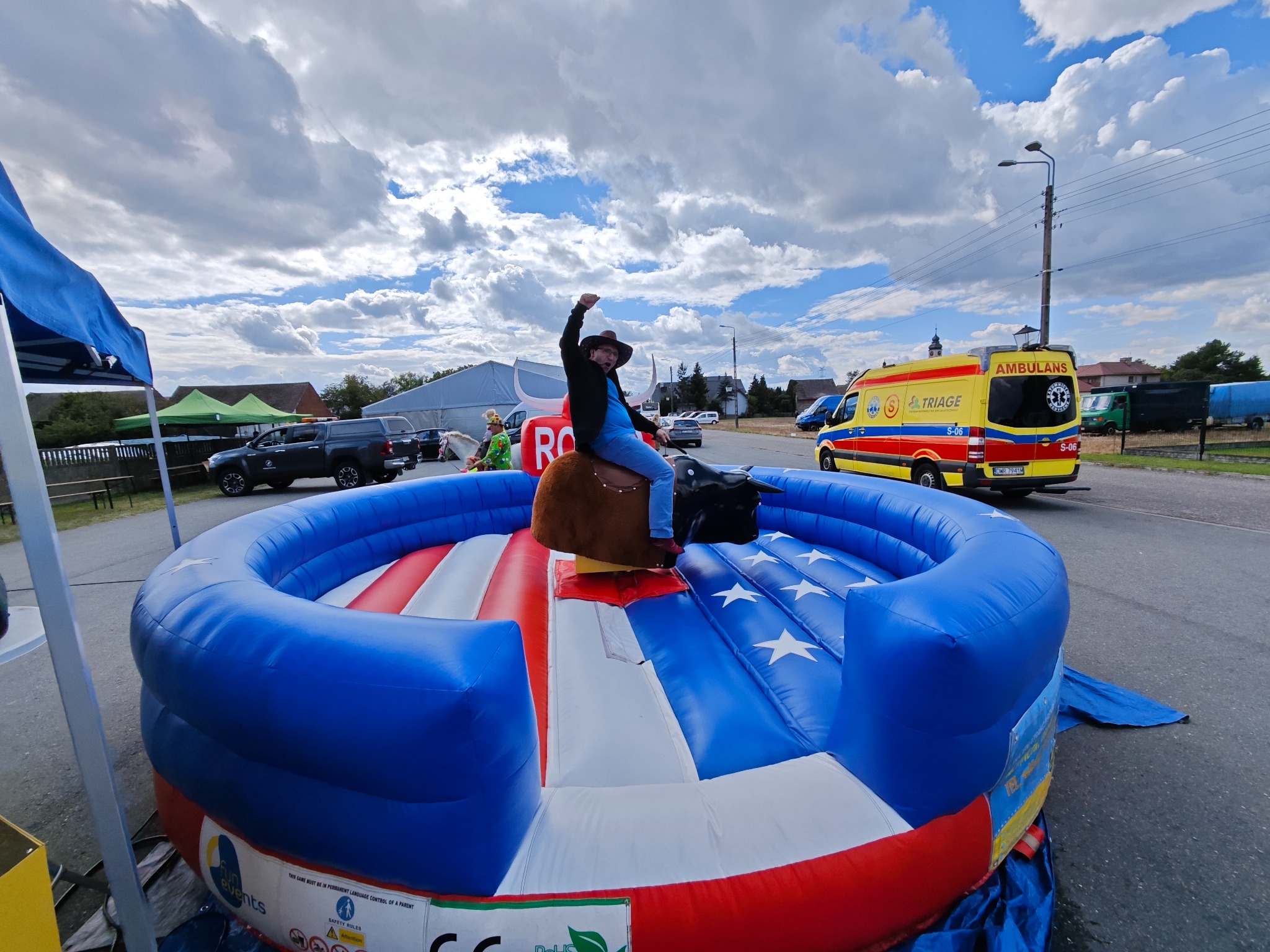 Mężczyzna w kapeluszu na dmuchanym byku rodeo, z podniesioną ręką w geście triumfu. W tle ambulans i inne pojazdy, częściowo zasłonięte przez dmuchany sprzęt.