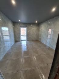 Puste pomieszczenie po remoncie z błyszczącą, beżową podłogą z płytek, szare ściany z efektem dekoracyjnym i szary sufit podwieszany z oświetleniem punktowym. Widoczne okno balkonowe.