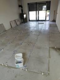 Szare płytki podłogowe układane na klej z użyciem krzyżyków dystansowych, widoczny fragment ściany i okna balkonowego, w tle piec.