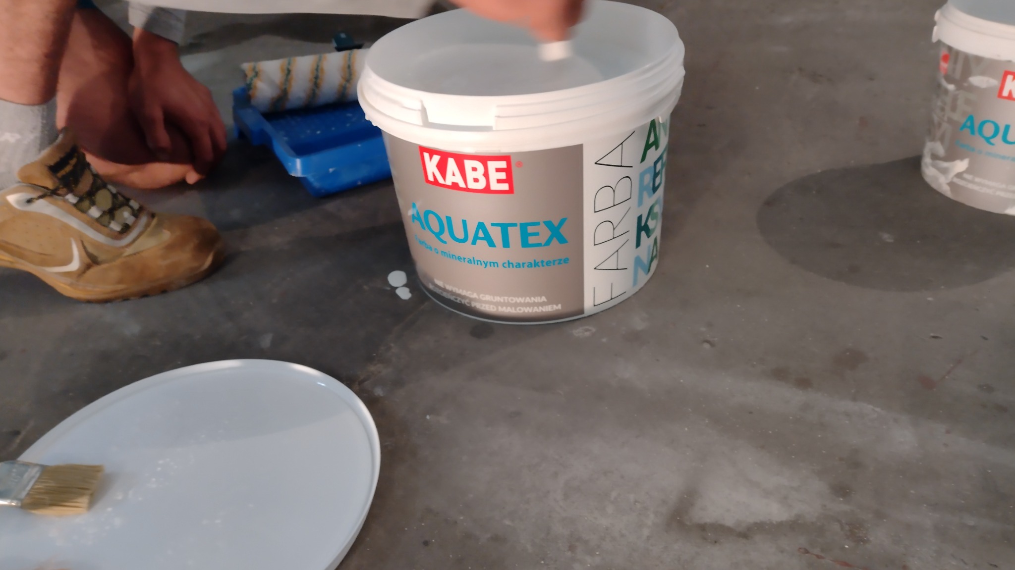 Przygotowanie do malowania: Wiadro z farbą KABE Aquatex, kuweta z wałkiem, pędzel na pokrywce i robocze buty na szarym podłożu.