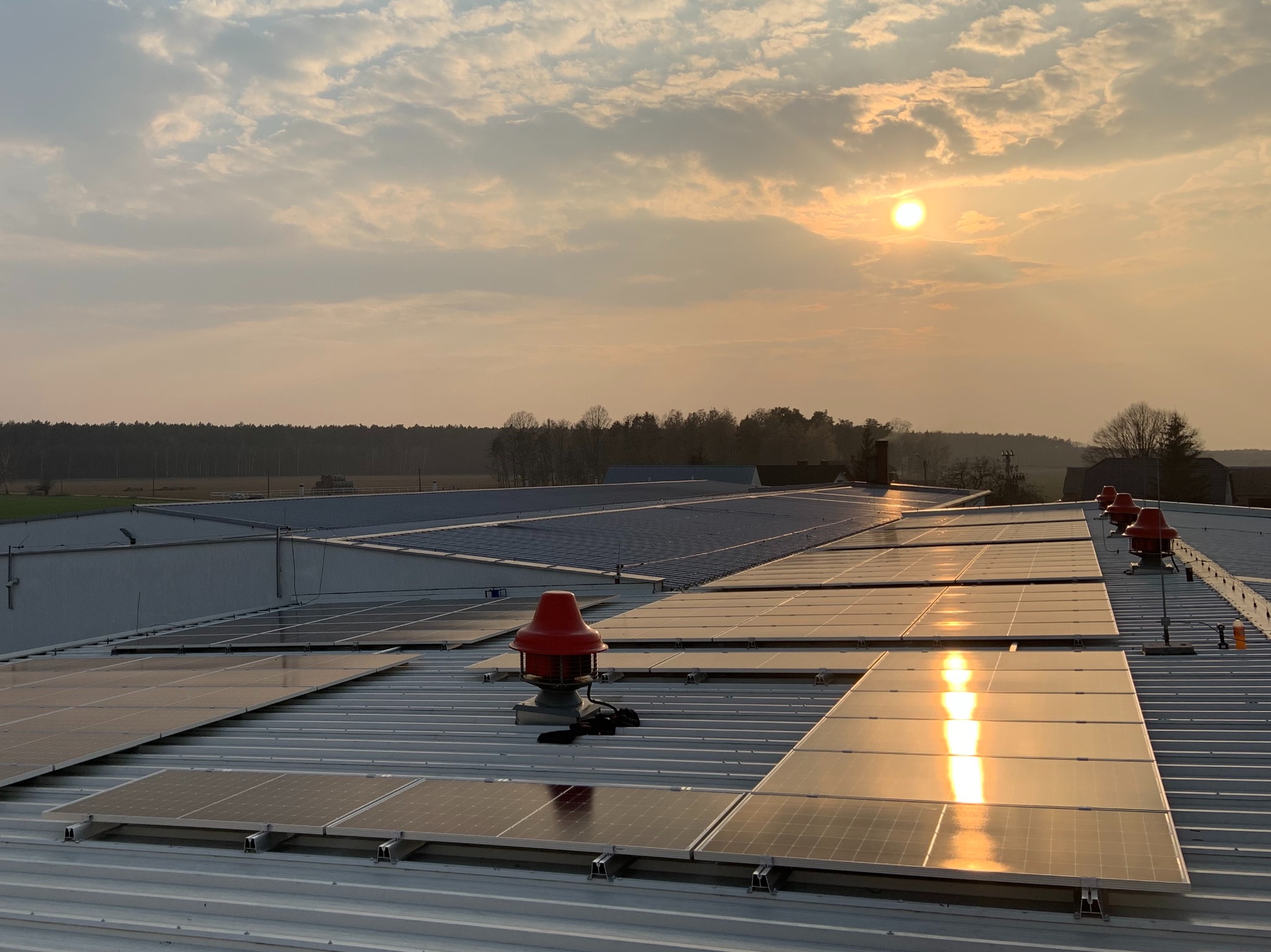 Instalacja paneli słonecznych na dachu hali przemysłowej oświetlona promieniami zachodzącego słońca, z widocznymi czerwonymi wentylatorami dachowymi.